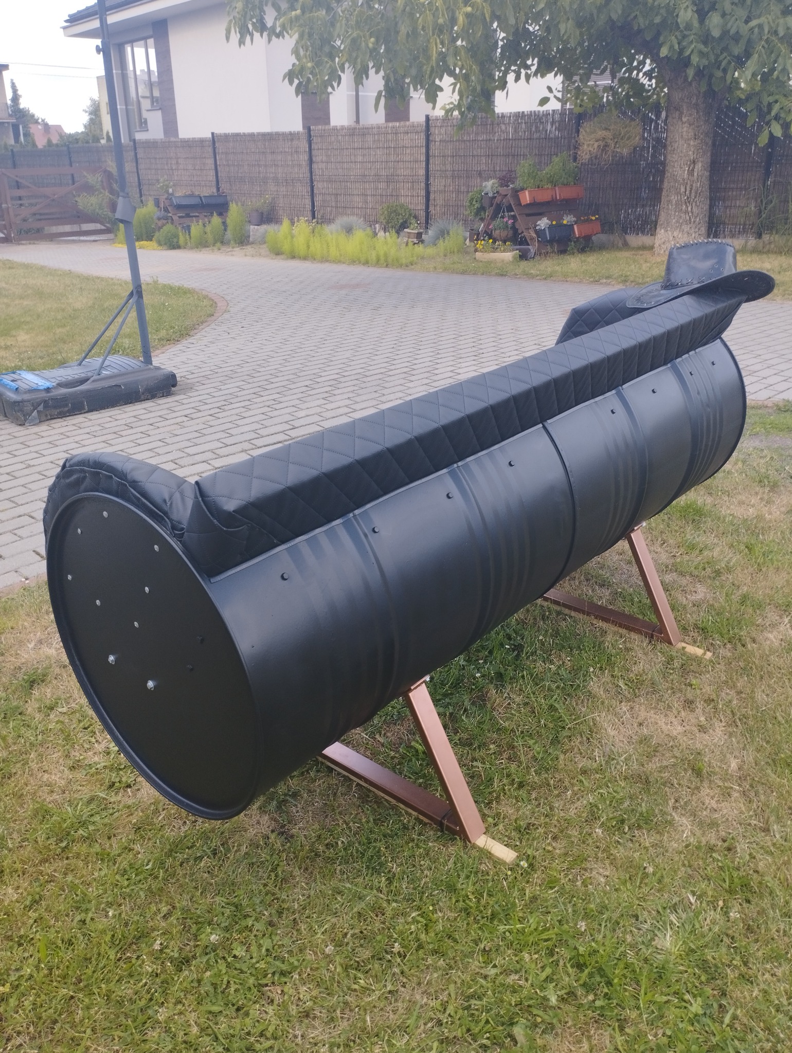 Czarna kanapa ogrodowa wykonana z metalowej beczki, z pikowanym siedziskiem, umieszczona na trawniku przed domem.