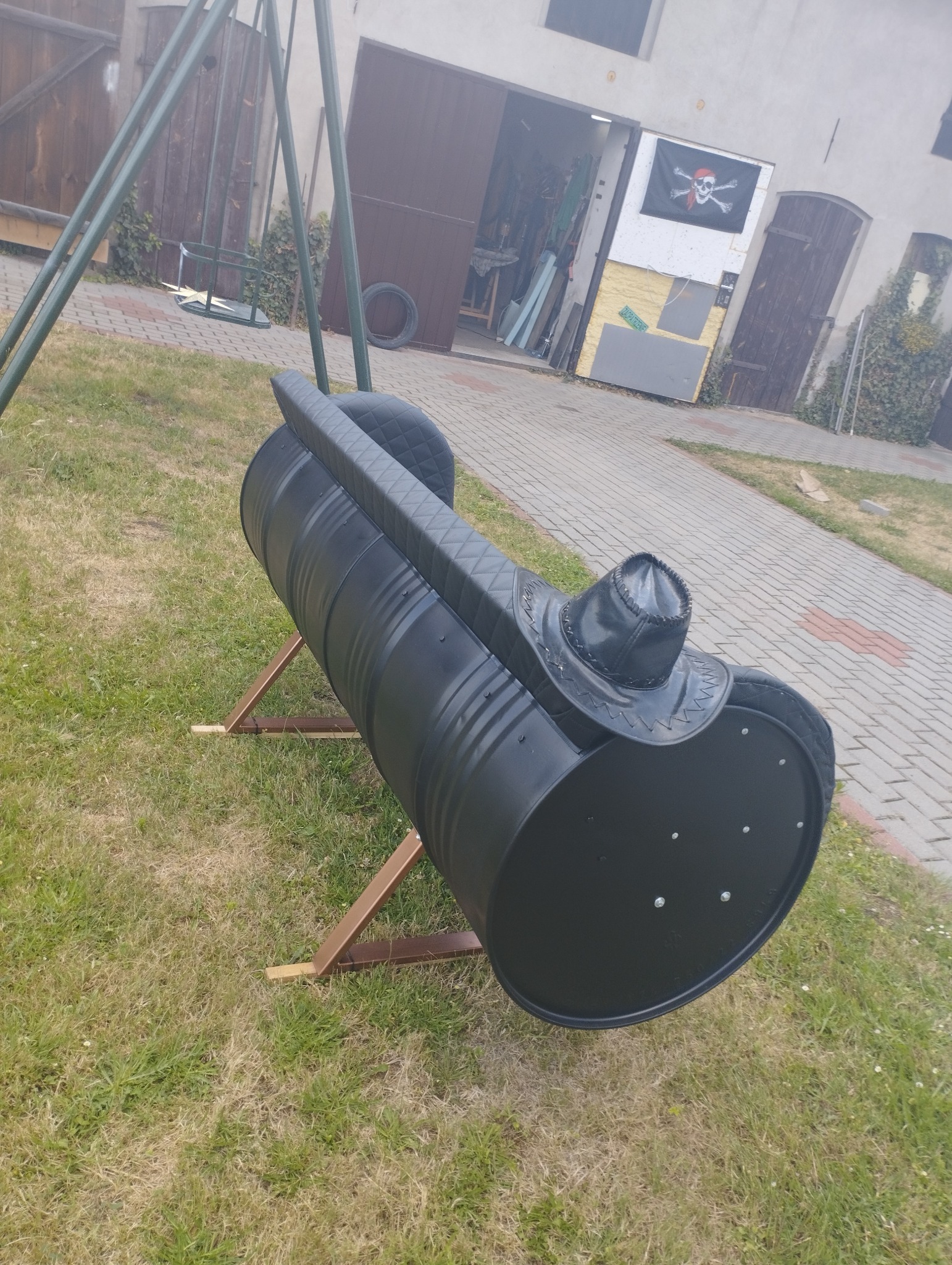 Czarny, metalowy bęben z siedziskiem i oparciem z pikowanej skóry ekologicznej, ustawiony na drewnianym stojaku na trawniku, z huśtawką i garażem w tle.