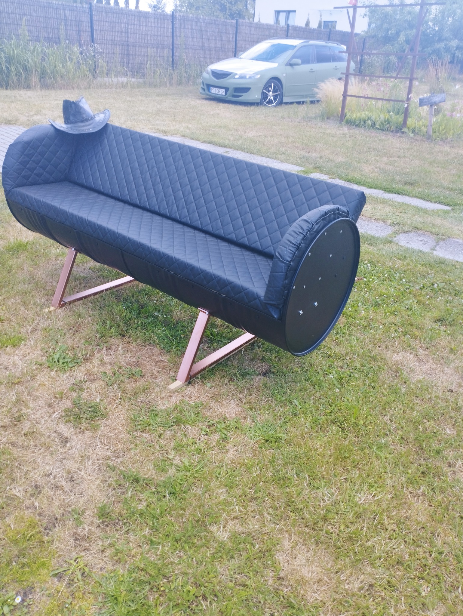 Czarna sofa wykonana z beczki, z pikowanym siedziskiem i oparciem, umieszczona na metalowych nogach w kolorze miedzianym, stoi na trawniku, z czarnym kapeluszem na oparciu i zielonym samochodem w tle.
