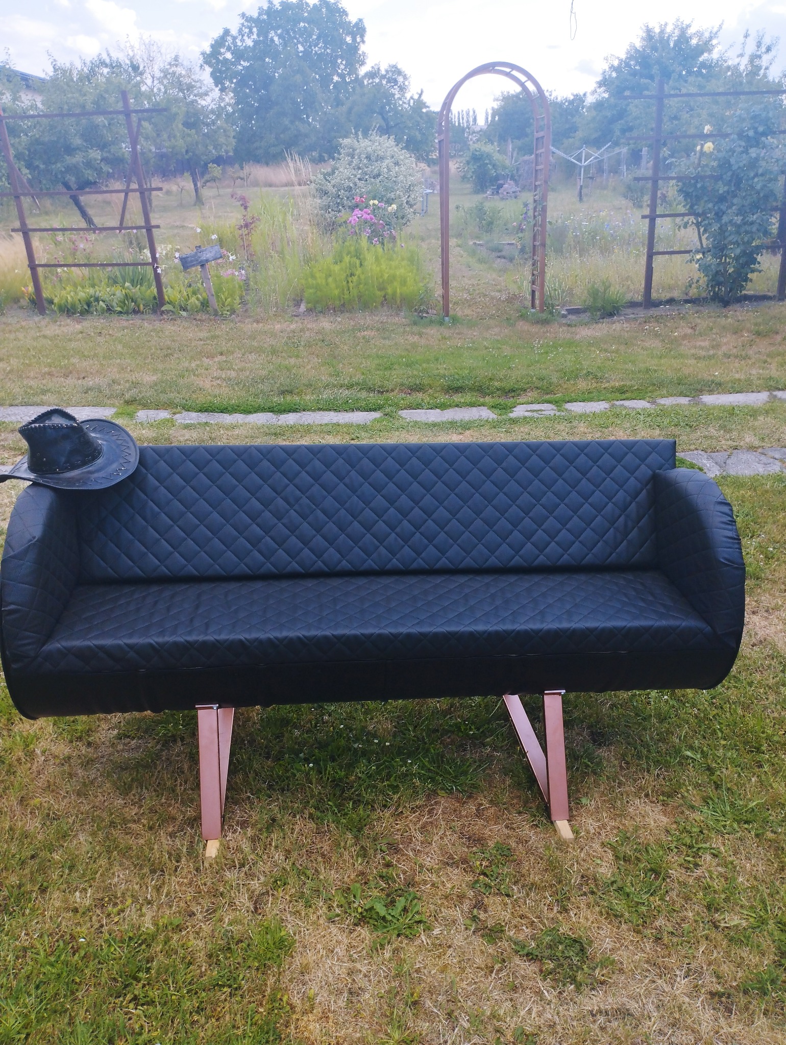 Czarna, pikowana sofa z ekoskóry na metalowych nogach w kolorze miedzi stoi na trawniku w ogrodzie, z czarnym kowbojskim kapeluszem na boku. W tle widać metalową konstrukcję i roślinność.