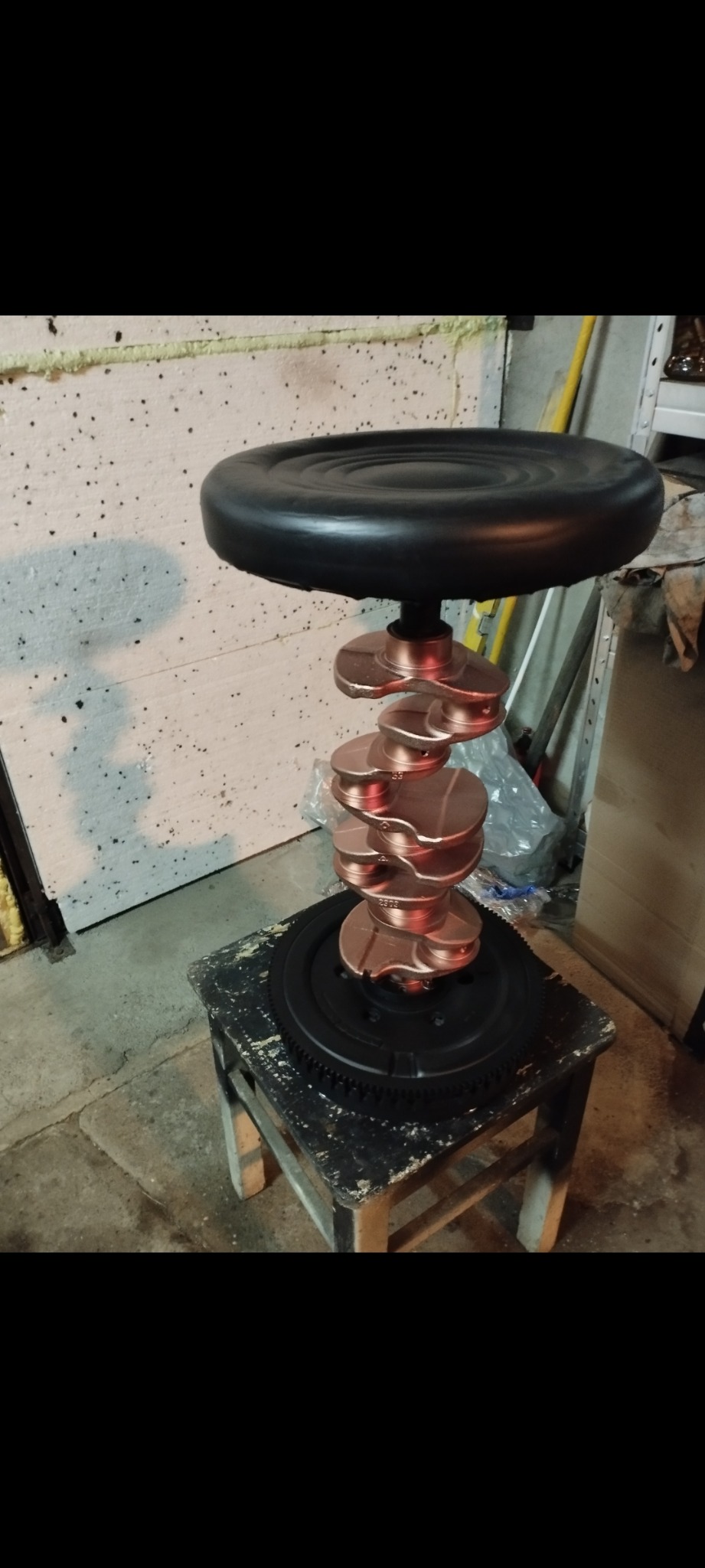 Taboret barowy wykonany z części samochodowych: czarnego siedziska, miedzianego wału korbowego i czarnego koła zamachowego, stojący na starym, pomalowanym stołku.