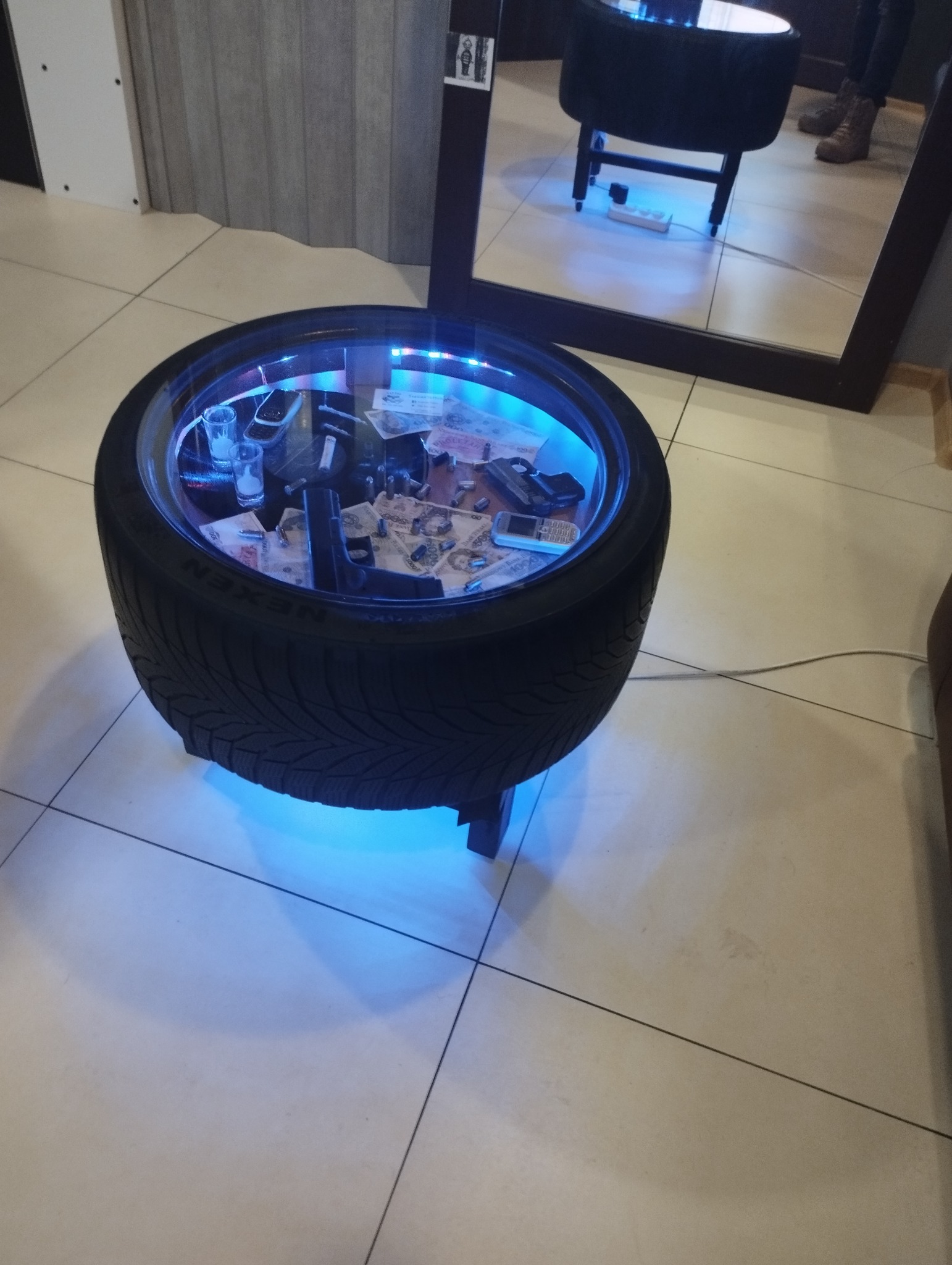 Stolik kawowy wykonany z opony samochodowej, z podświetleniem LED i szklanym blatem, wypełniony rekwizytami imitującymi broń, pieniądze i telefony komórkowe, odbity w lustrze.