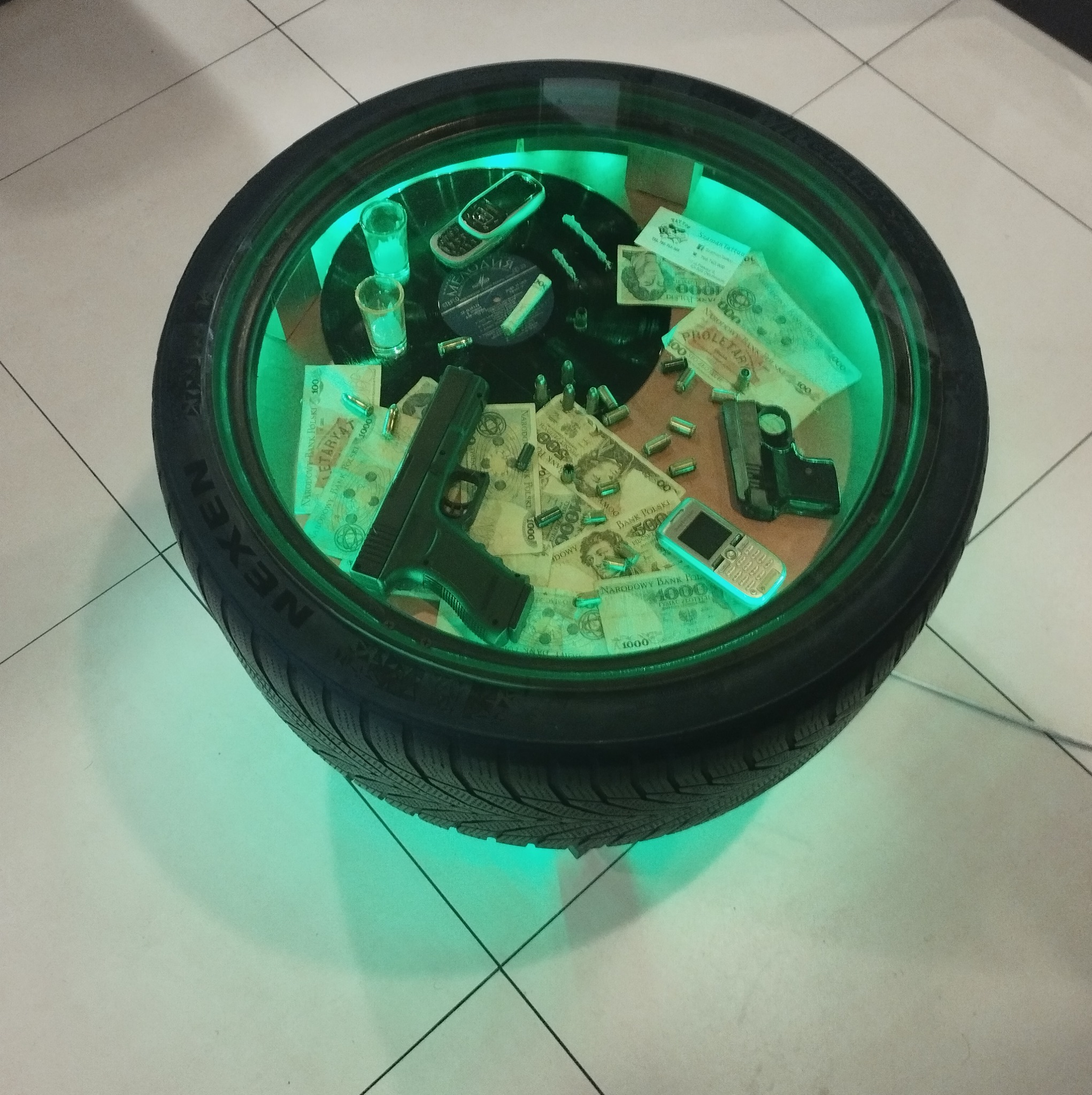 Stolik kawowy wykonany z opony samochodowej, podświetlony na zielono, z szybą na górze, na której leżą banknoty, naboje, telefony komórkowe i dwa kieliszki