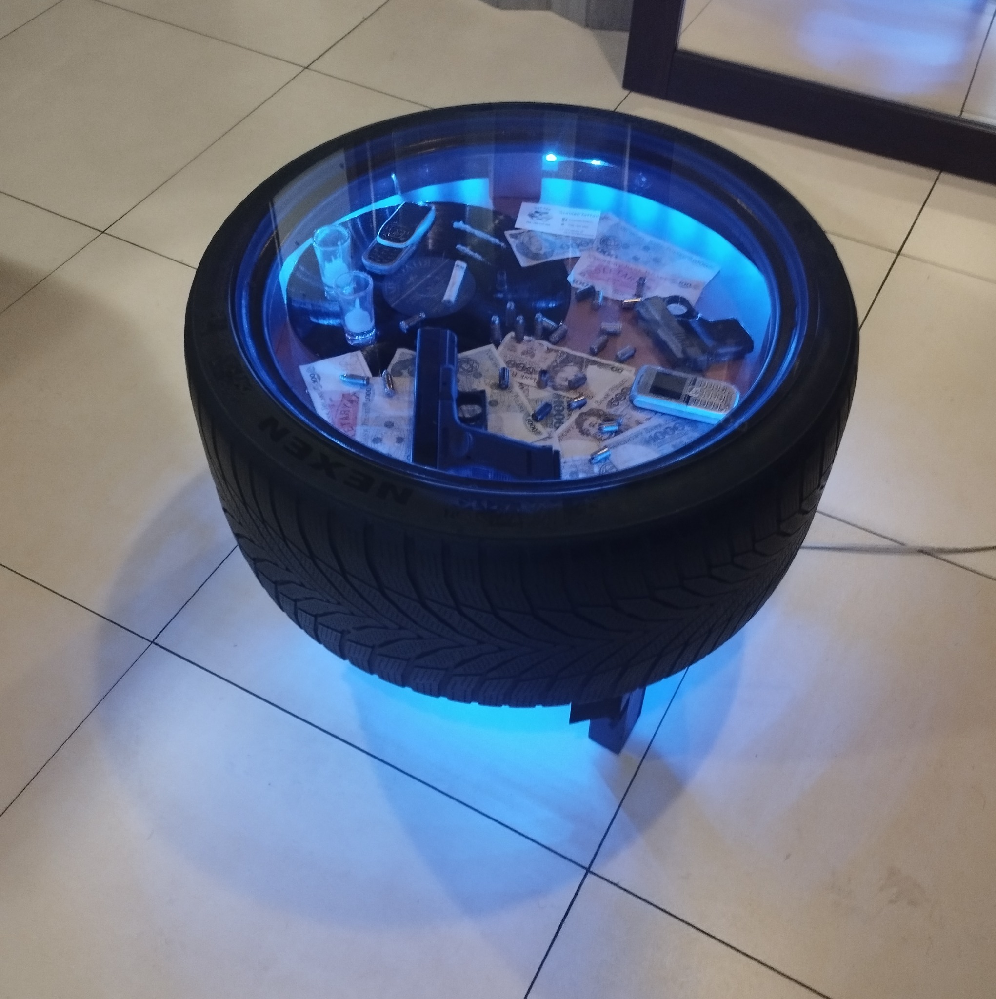 Stolik kawowy wykonany z opony samochodowej, podświetlany na niebiesko, z szybą, wewnątrz którego znajdują się banknoty, naboje, pistolet i stare telefony komórkowe.
