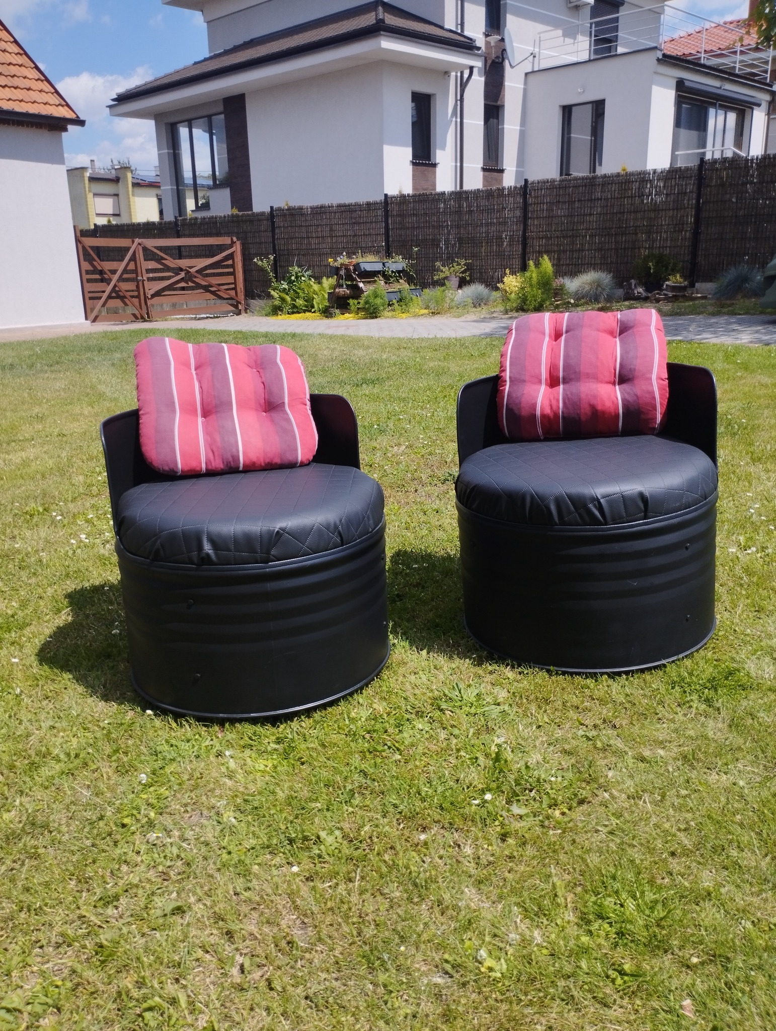 Dwa czarne fotele ogrodowe wykonane z beczek z pikowanymi siedziskami i poduszkami w czerwono-białe paski, stojące na trawniku przed nowoczesnym domem z drewnianym ogrodzeniem.