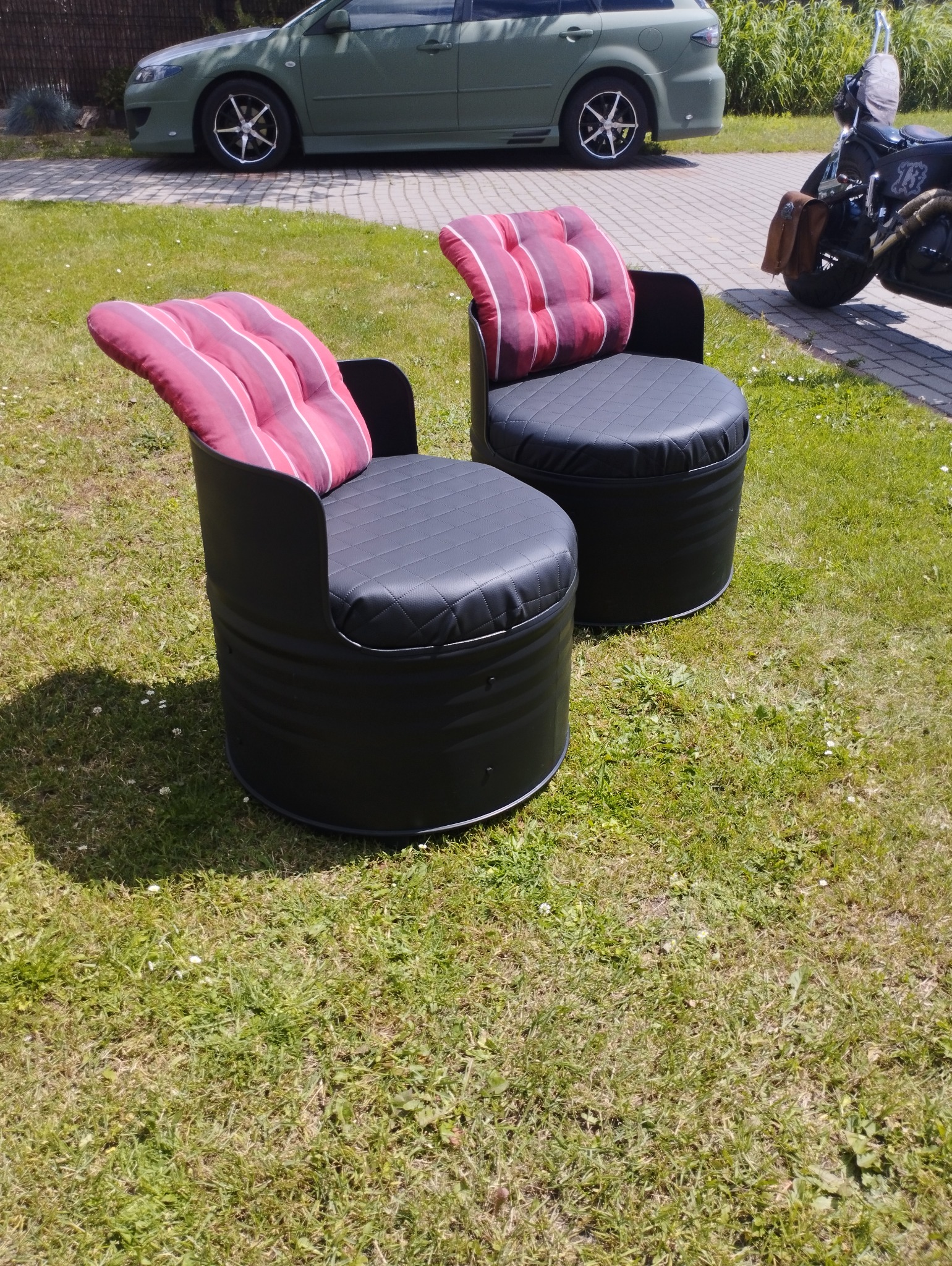 Dwa czarne fotele wykonane z beczek z pikowanymi siedziskami i oparciami z czerwono-białego materiału w paski, stojące na trawniku, w tle zaparkowany samochód i motocykl.