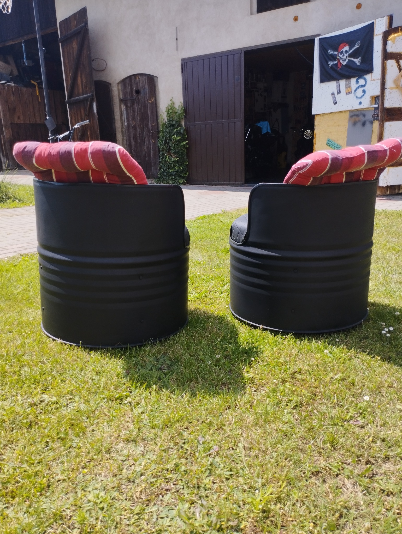 Dwa czarne fotele wykonane z metalowych beczek z czerwono-białymi poduszkami, stojące na trawniku przed otwartymi drzwiami garażu z wywieszoną piracką flagą.