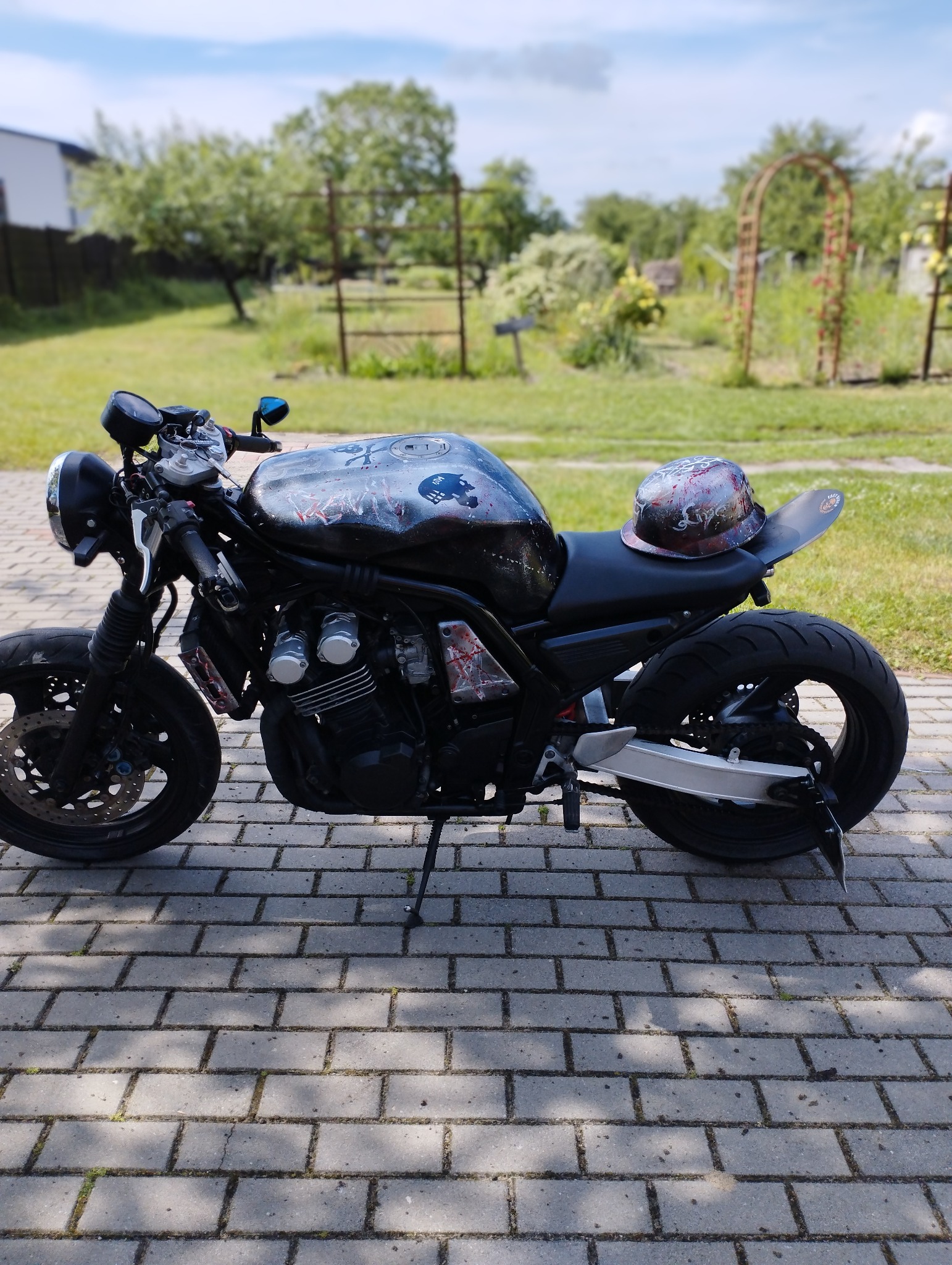Motocykl typu cafe racer z niestandardowym malowaniem czaszek na baku i kasku, zaparkowany na brukowanej ścieżce w ogrodzie z dekoracyjnymi łukami.