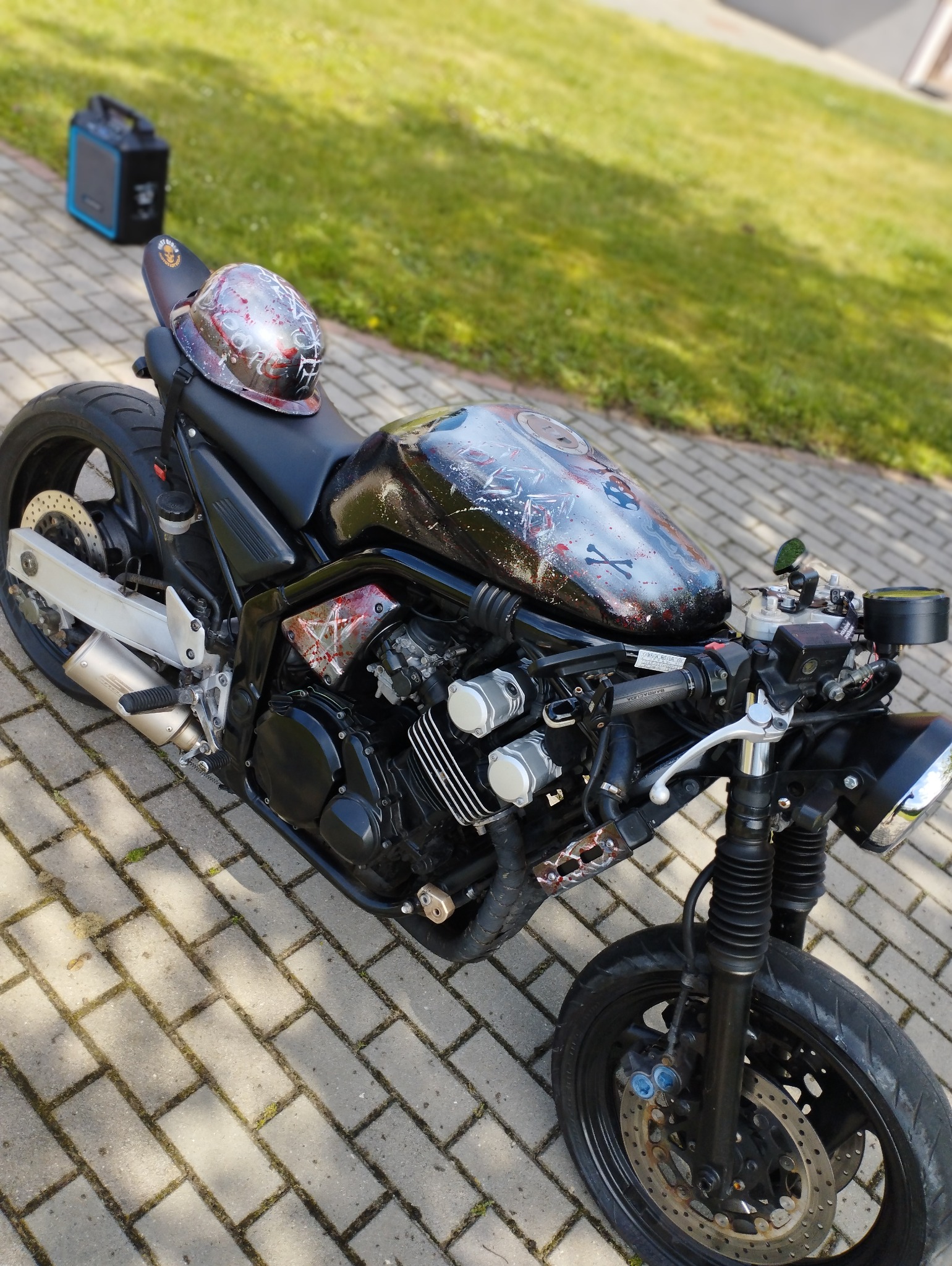 Czarny motocykl typu cafe racer z customowym malowaniem czaszek i kości na baku, obok leży kask z podobnym motywem malarskim, w tle przenośna stacja zasilania.