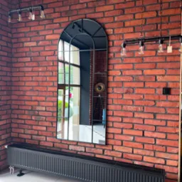 Loftowe wnętrze w Pile z czerwoną cegłą na ścianie, grzejnikiem panelowym i wysokim, łukowym lustrem z czarną ramą, w którym odbija się lampa na trójnogu.