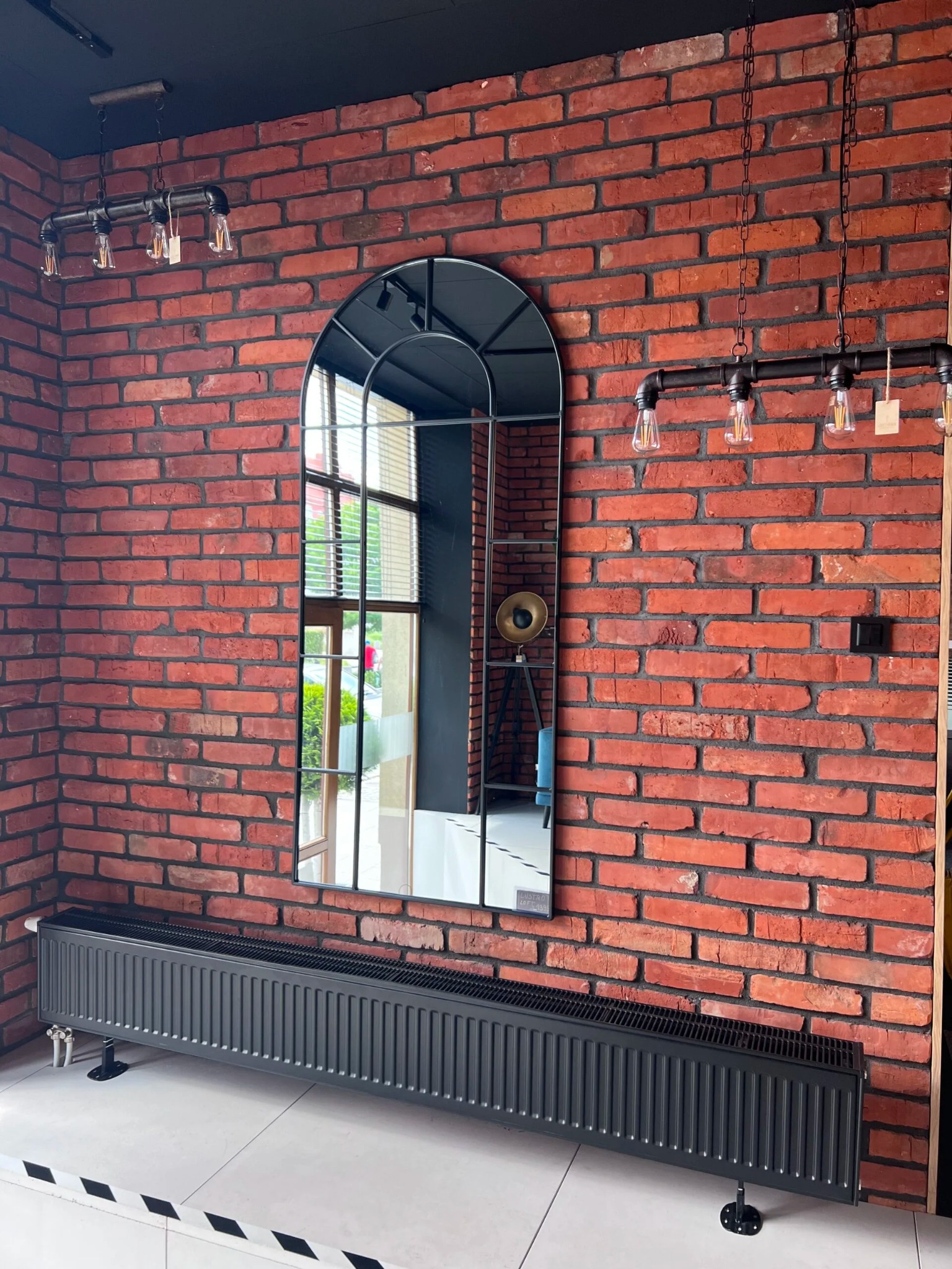 Loftowe wnętrze w Pile z czerwoną cegłą na ścianie, grzejnikiem panelowym i wysokim, łukowym lustrem z czarną ramą, w którym odbija się lampa na trójnogu.