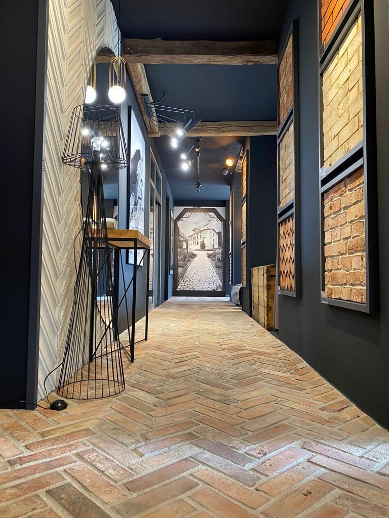Długi korytarz z podłogą z cegły ułożonej w jodełkę, oświetlony nowoczesnymi lampami i halogenami, z dekoracyjnymi panelami ściennymi i obrazem perspektywicznym na końcu.