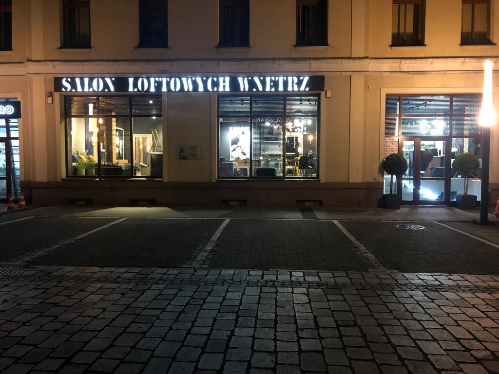 Fasada budynku z napisem 'SALON LOFTOWYCH WNĘTRZ' nad trzema dużymi oknami wystawowymi, widok z zewnątrz na brukowany parking nocą.