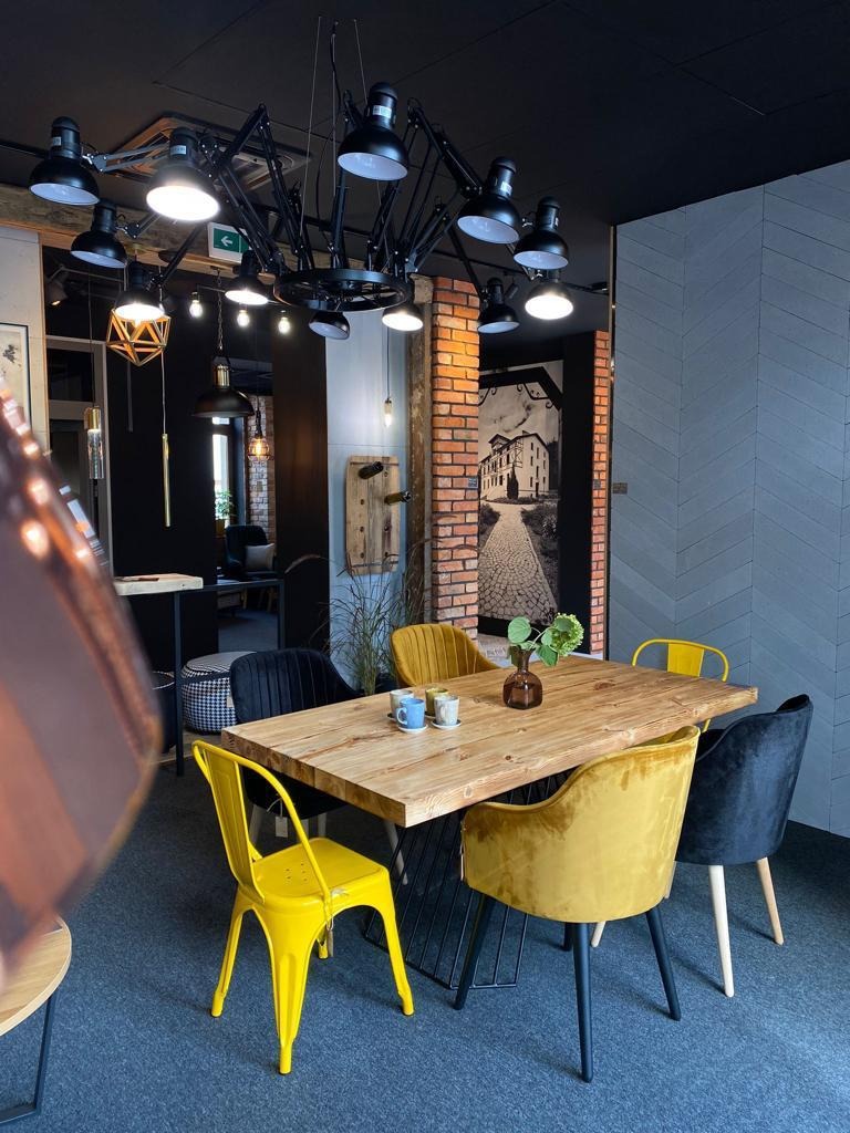 Jadalnia z drewnianym stołem na metalowej podstawie, otoczonym krzesłami w kolorach żółtym i czarnym, oświetlona industrialną lampą sufitową, ściana z cegły i szara ściana z wzorem w jodełkę,...