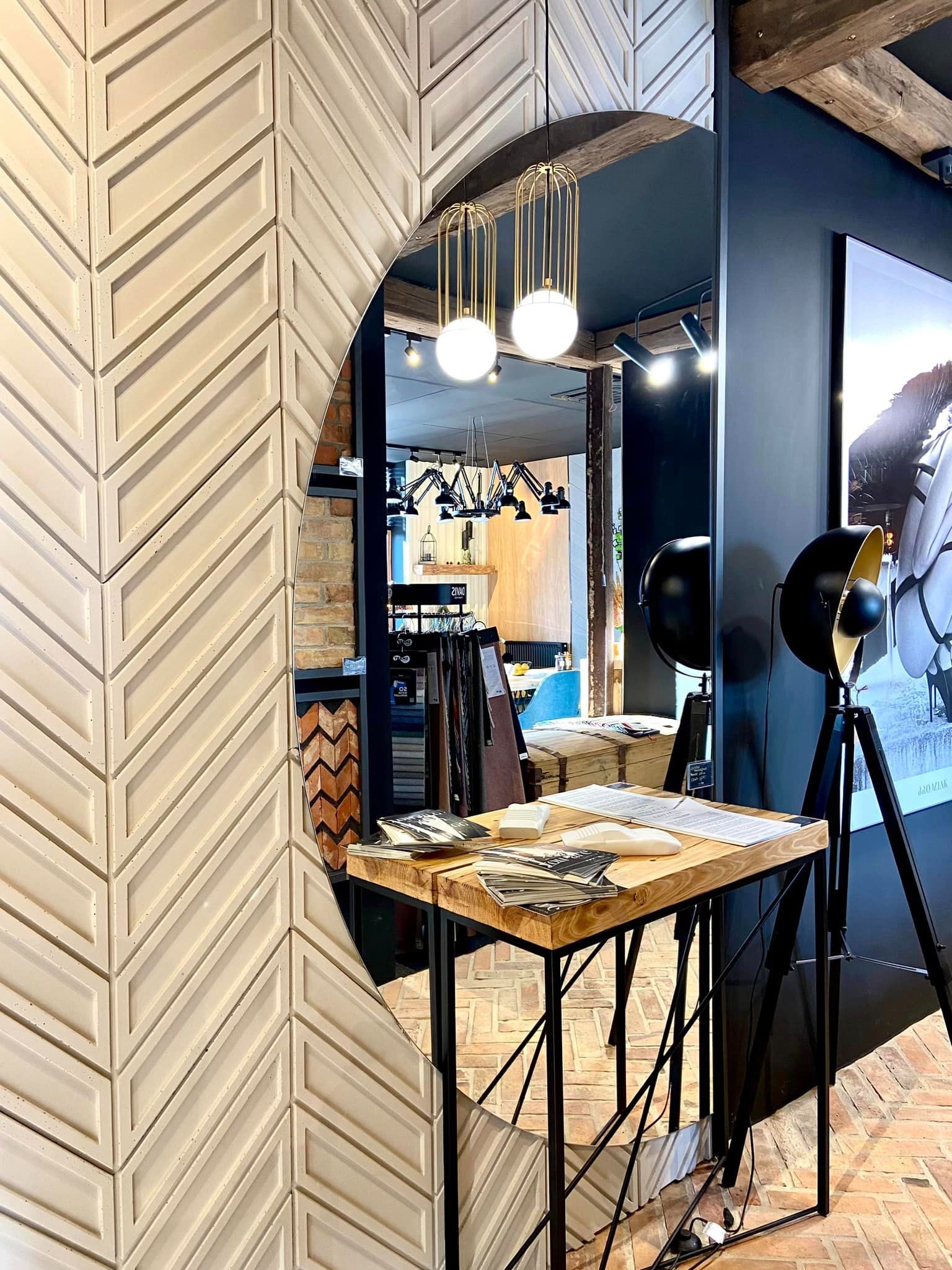 Wystrój wnętrza sklepu z dekoracyjną ścianą w jodełkę, owalnym lustrem i lampami wiszącymi w złotych klatkach, prezentacja materiałów na drewnianym stole z metalową podstawą.