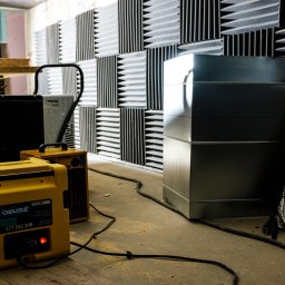 OSUSZ GDAŃSK SPÓŁKA Z OGRANICZONĄ ODPOWIEDZIALNOŚCIĄ - Żółty osuszacz powietrza z włączoną czerwoną diodą, stojący na drewnianej desce w pomieszczeniu z panelami akustycznymi i metalową konstrukcją. Widoczny kabel zasilający.