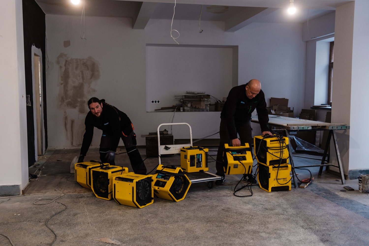 Dwóch mężczyzn w czarnych uniformach rozstawia żółte osuszacze powietrza TROTEC w surowym, remontowanym pomieszczeniu z odsłoniętymi ścianami i betonową podłogą.