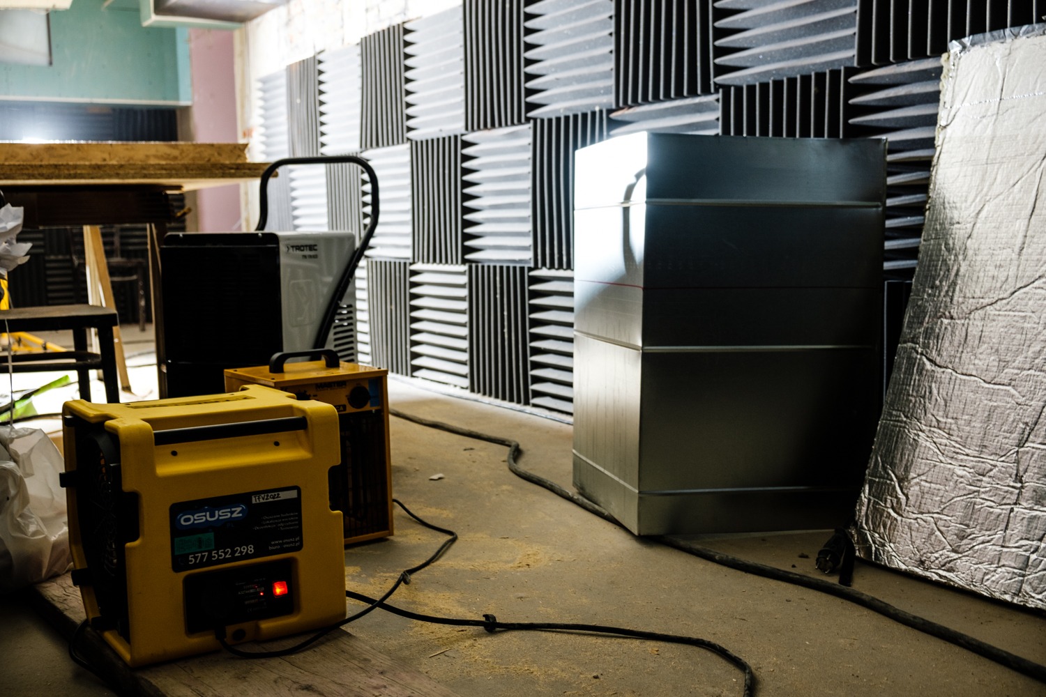 Żółty osuszacz powietrza z włączoną czerwoną diodą, stojący na drewnianej desce w pomieszczeniu z panelami akustycznymi i metalową konstrukcją. Widoczny kabel zasilający.