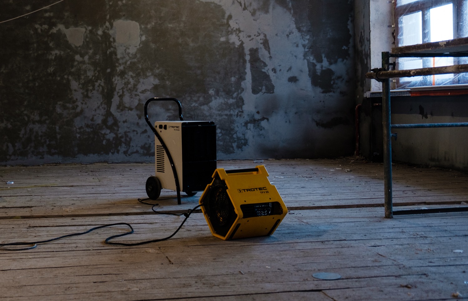 Dwa urządzenia Trotec na drewnianej podłodze w surowym, remontowanym pomieszczeniu: biało-czarny osuszacz z rączką i żółta turbina susząca Trotec TFV 29, kabel na podłodze, ściany z widocznymi...