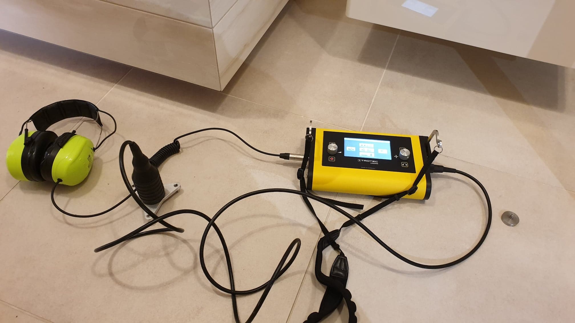Żółty miernik wilgotności Trotec LD6000 z podłączonymi przewodami i sondą, obok leżą ochronne słuchawki na tle beżowych płytek podłogowych i fragmentu białej szafki.