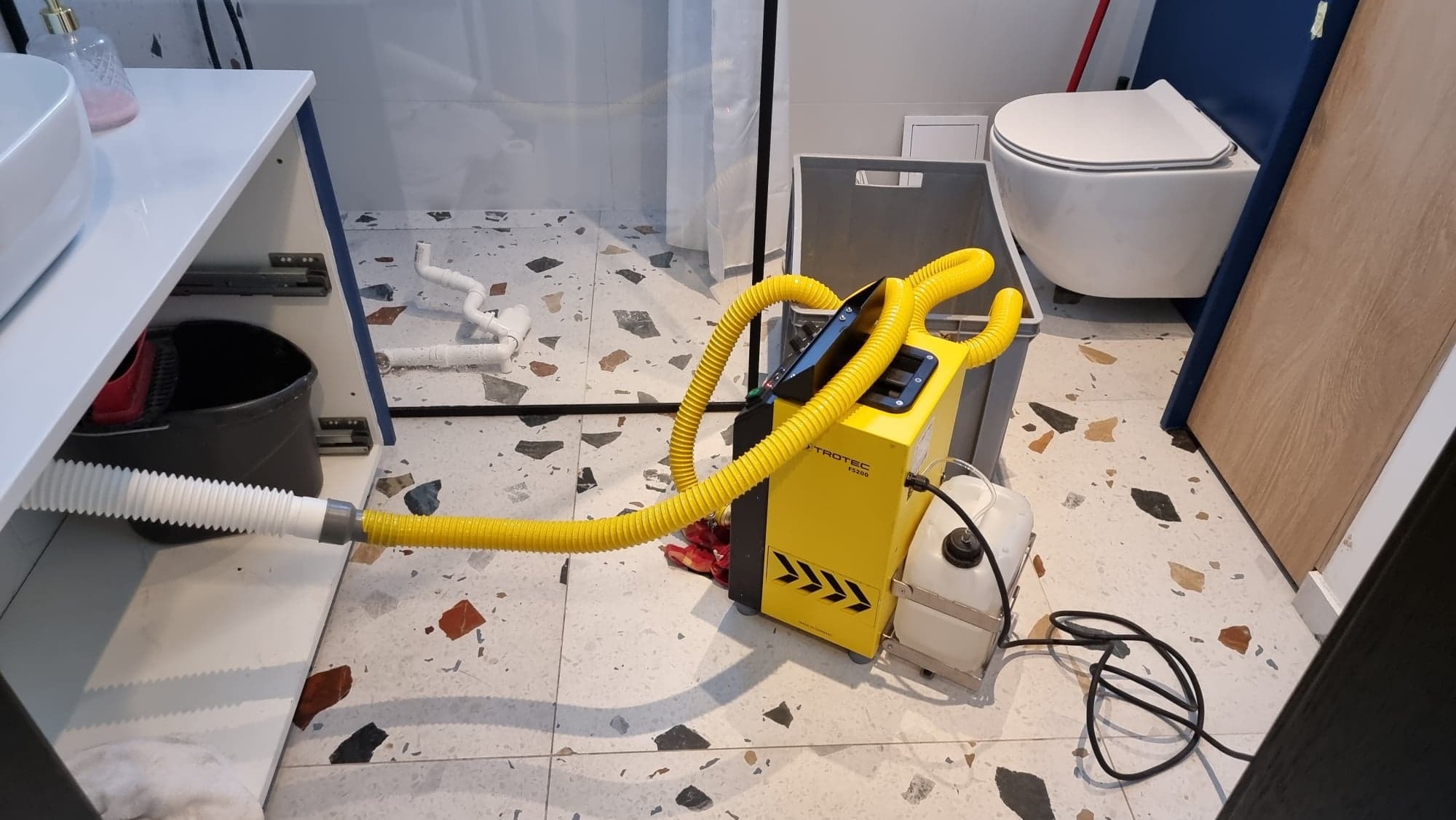 Żółty osuszacz Trotec PS200 z podłączonymi wężami, ustawiony w łazience z mozaikową podłogą, widok na umywalkę i toaletę.