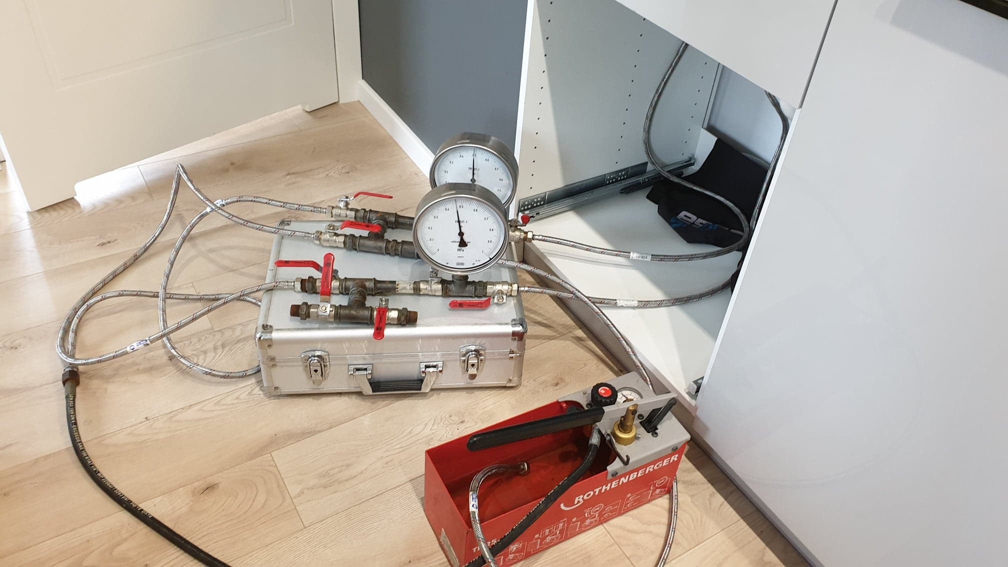 Urządzenie do pomiaru ciśnienia Rothenberger z manometrami, zaworami i przewodami, ustawione na podłodze obok otwartej szafki kuchennej, podczas testu szczelności instalacji.