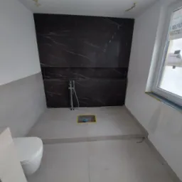 Wnętrze łazienki w trakcie remontu, widoczna zabudowa z czarnej płyty imitującej marmur, biała toaleta oraz przygotowane miejsce na prysznic z odpływem liniowym.