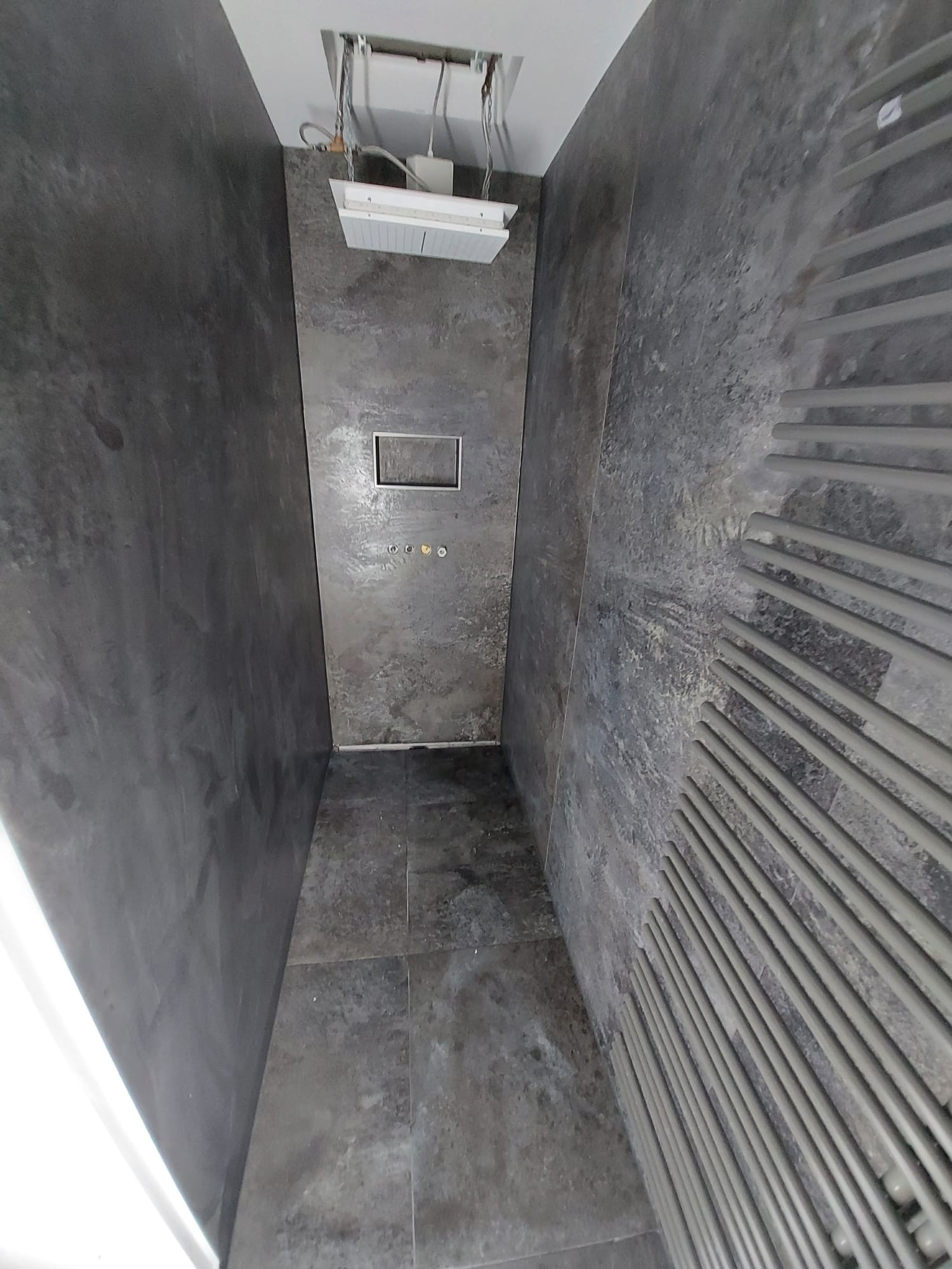 Nowoczesna łazienka z ciemnoszarą, teksturowaną okładziną ścian i podłogi, widoczny grzejnik drabinkowy, panel sufitowy z oświetleniem i instalacją.