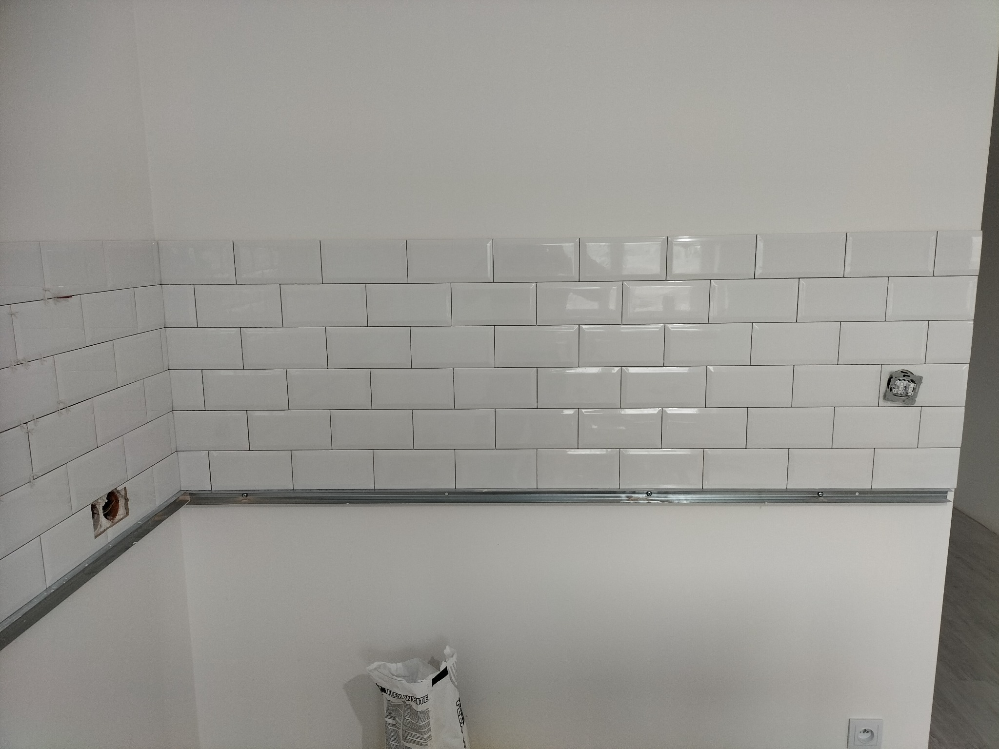 Świeżo ułożone białe, prostokątne płytki na ścianie, z metalową listwą poziomującą i workiem zaprawy klejowej u dołu, widoczne puszki elektryczne bez osprzętu.