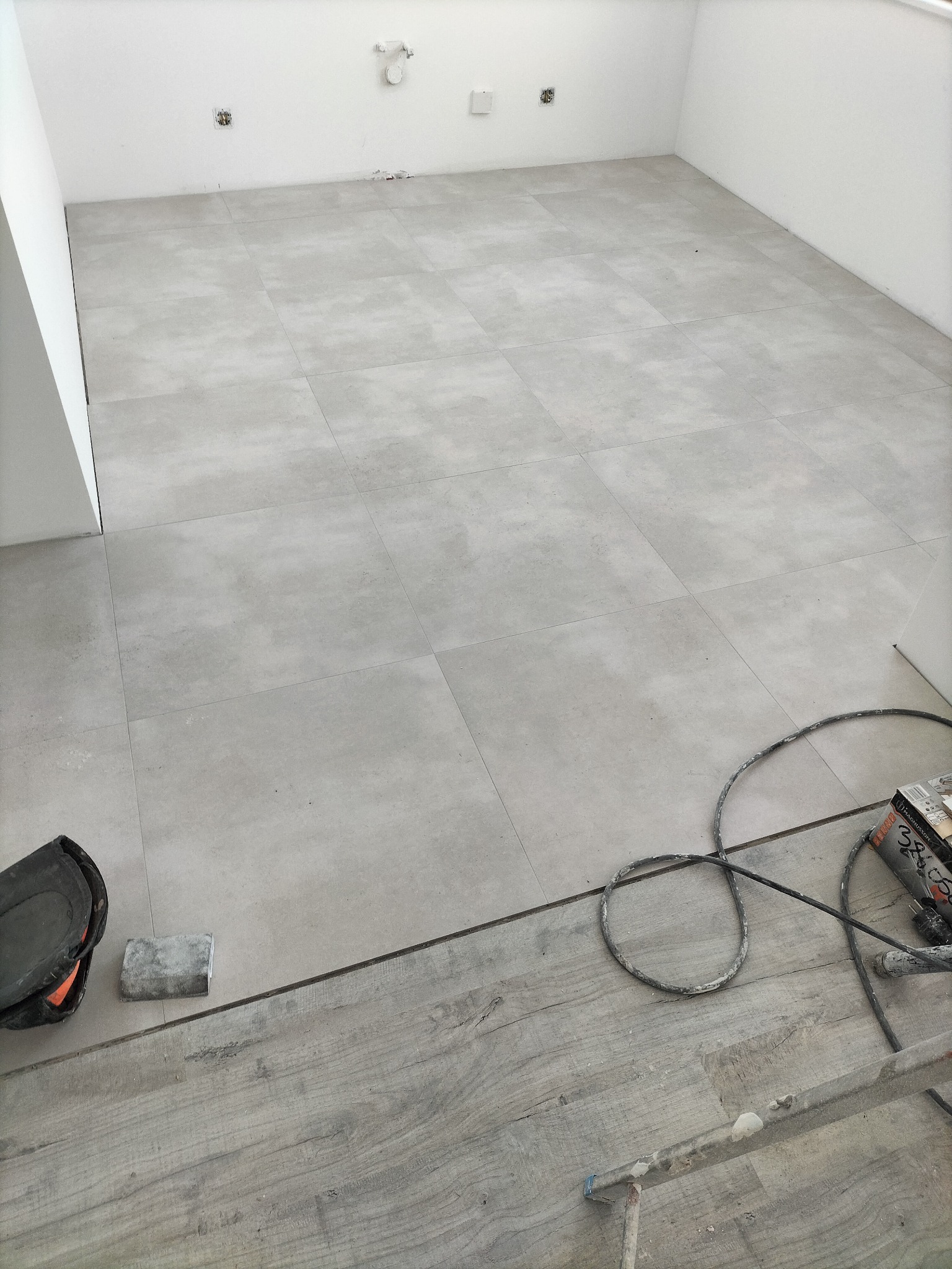 Ułożona jasnoszara płytka podłogowa w łazience, widoczne narzędzia glazurnicze i fragment drewnianej podłogi przed wejściem do pomieszczenia.