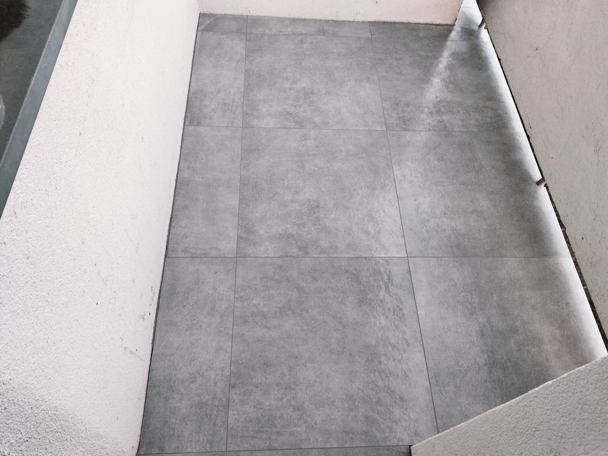 Szare płytki podłogowe ułożone w kwadraty, widoczne fugi, ściana z szorstką fakturą. Ujęcie z góry, widoczne krawędzie ścian. Naturalne światło.