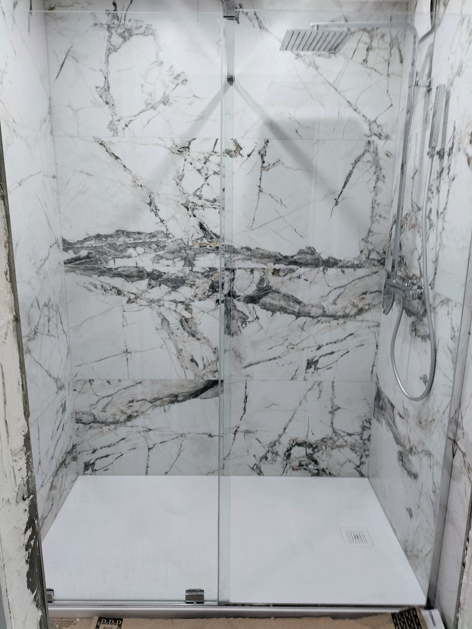 Kabina prysznicowa z deszczownicą, wykończona płytkami imitującymi marmur. Widoczne białe brodzik i szklane drzwi przesuwne. Częściowo wykończone ściany.