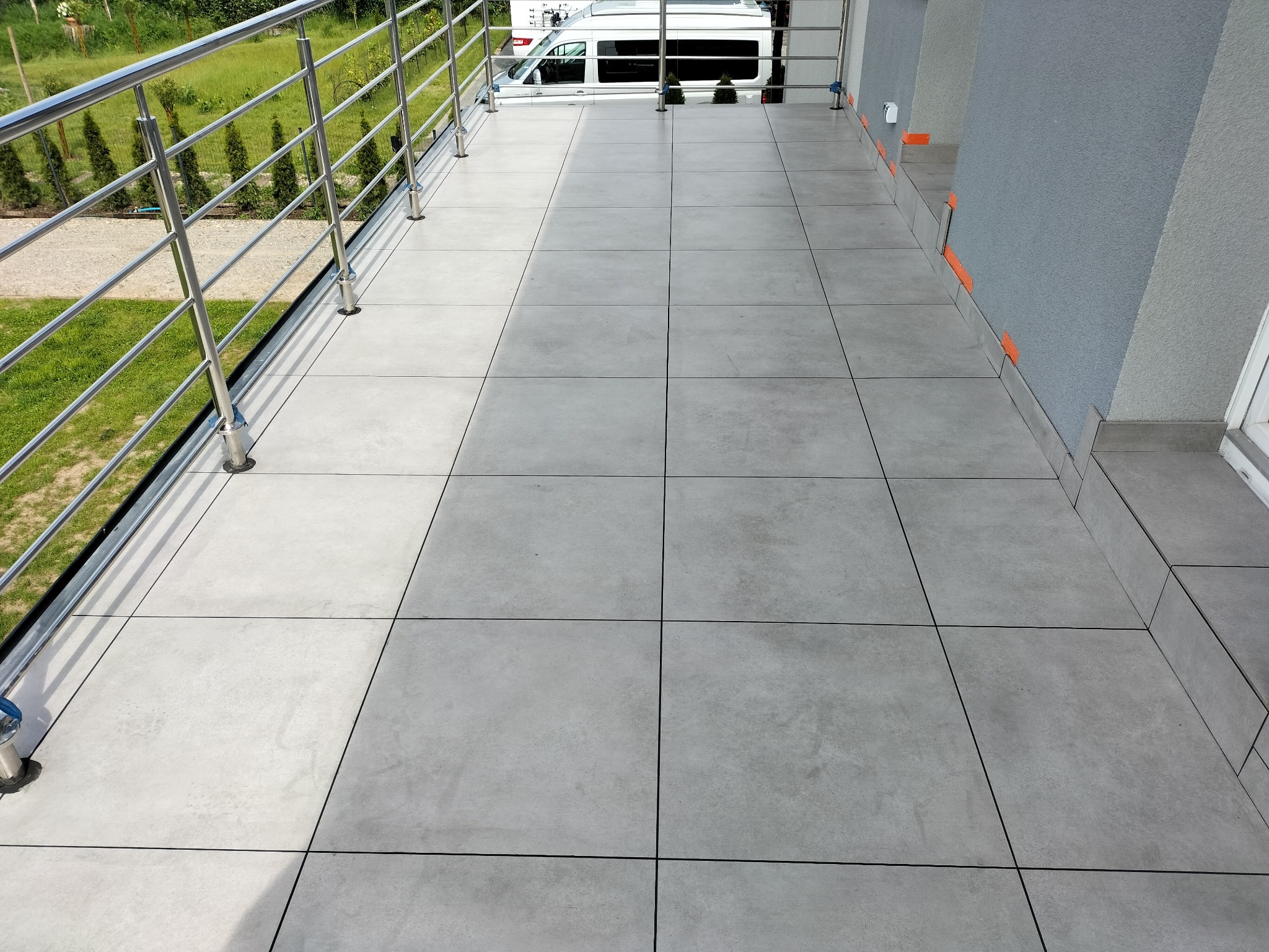 Wyłożony szarymi płytkami balkon z metalową balustradą. Widoczne pomarańczowe elementy dystansowe przy ścianie. W tle zieleń i zaparkowany biały samochód.