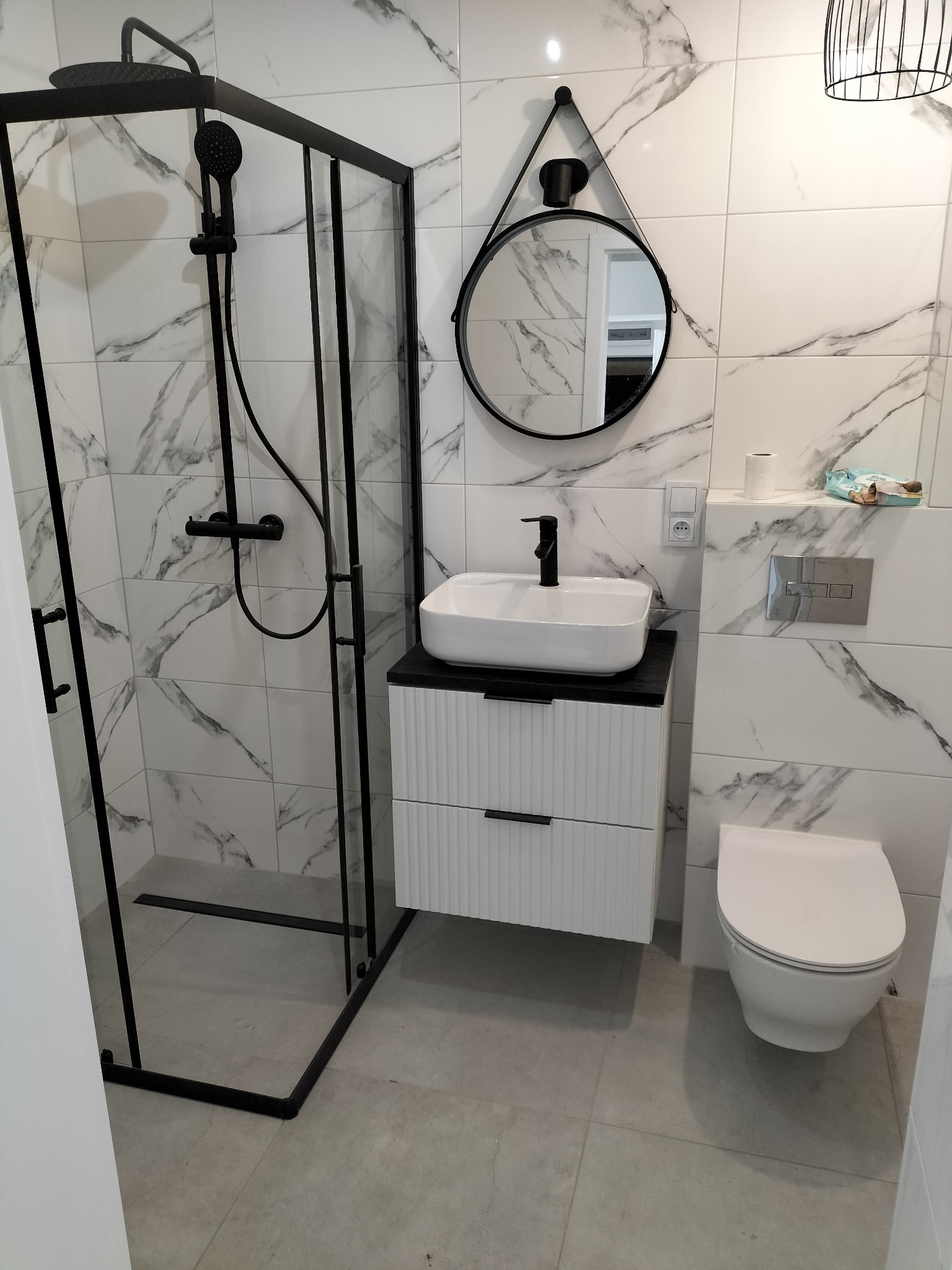 Nowoczesna łazienka z kabiną prysznicową typu walk-in z czarnymi profilami, umywalką nablatową i płytkami imitującymi marmur. Minimalistyczny design.