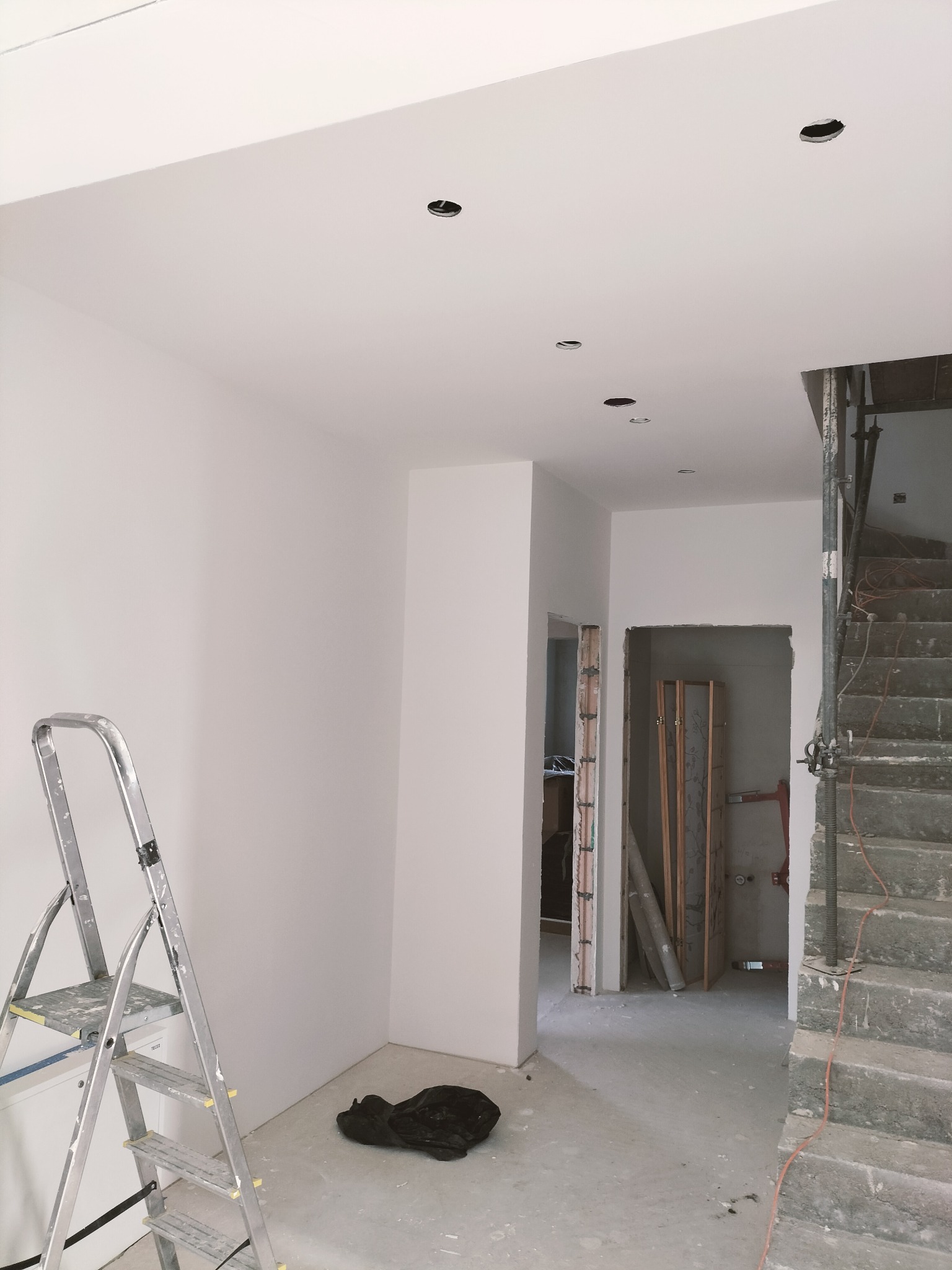 Wnętrze w trakcie wykończenia: białe ściany, sufit z otworami na oświetlenie, drabina malarska, surowe schody i widoczne przygotowania do montażu drzwi.