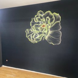 TarTapety - Czarna ściana z ręcznie malowanym, świecącym motywem kwiatowym, widoczny fragment podłogi z paneli i nowoczesna lampa sufitowa.