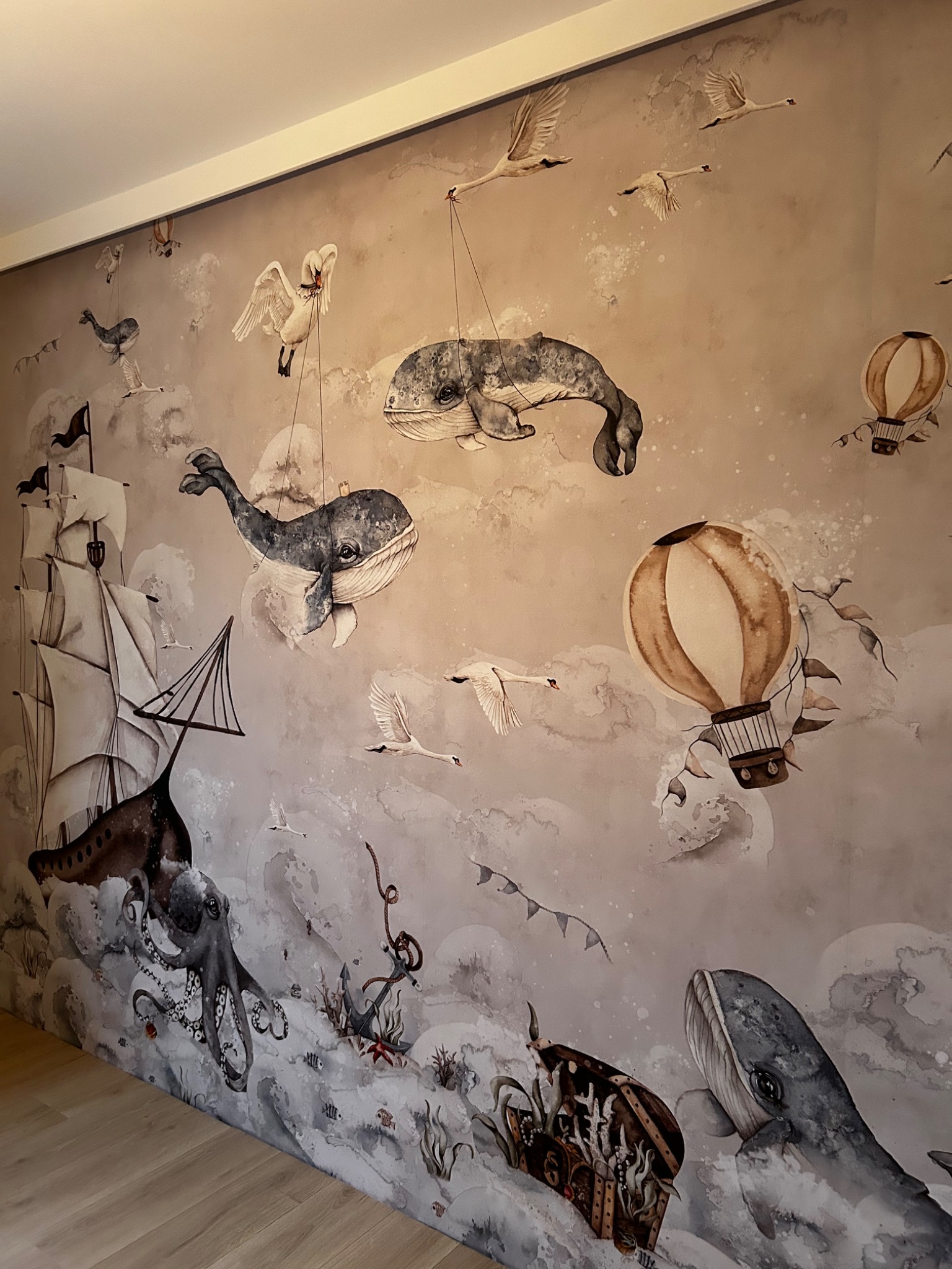 Dziecięca ściana z fototapetą przedstawiającą motywy morskie, w tym statek, ośmiornicę, wieloryby i balony na tle chmur. Tapeta w odcieniach szarości i beżu.