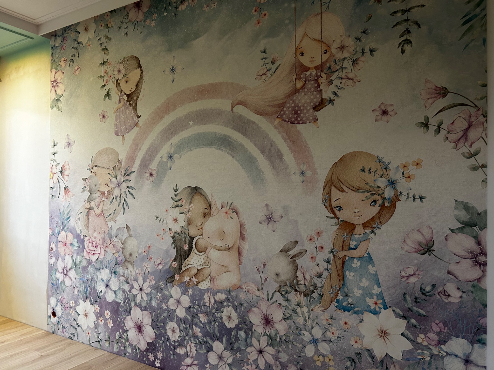 Ściana w pokoju dziecięcym z fototapetą przedstawiającą ilustracje dziewczynek, zwierząt i kwiatów w pastelowych kolorach, z motywem tęczy w tle.
