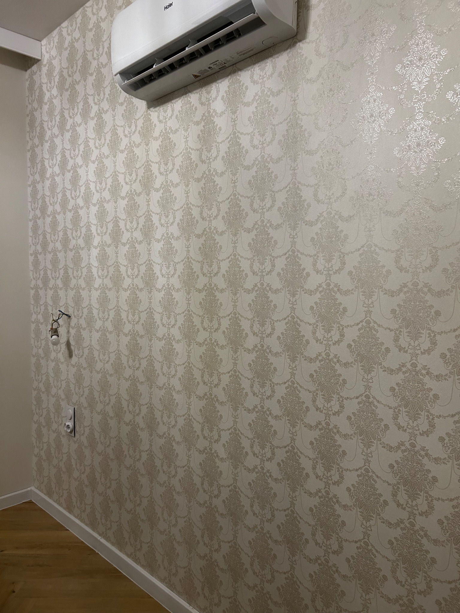 Ściana pokryta beżową tapetą w klasyczny wzór, widoczny klimatyzator Haier, gniazdko elektryczne oraz fragment instalacji elektrycznej z żarówką.