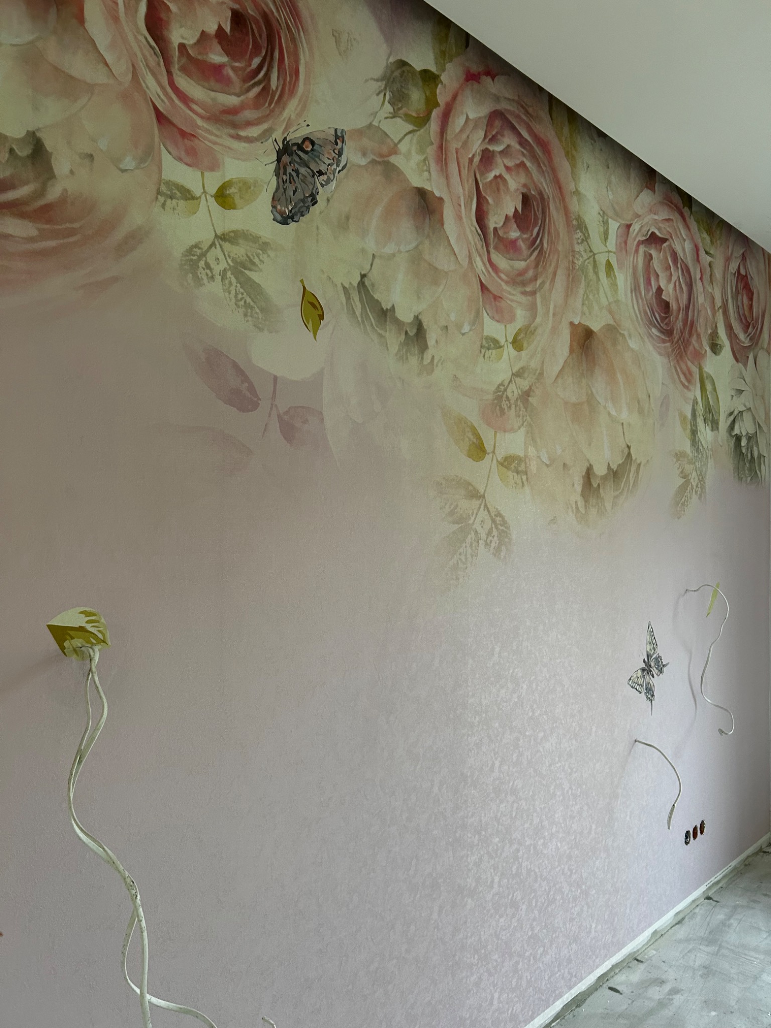 Ściana pokryta tapetą z motywem róż i motyli, widoczne niedokończone prace elektryczne przy suficie i gniazdkach.