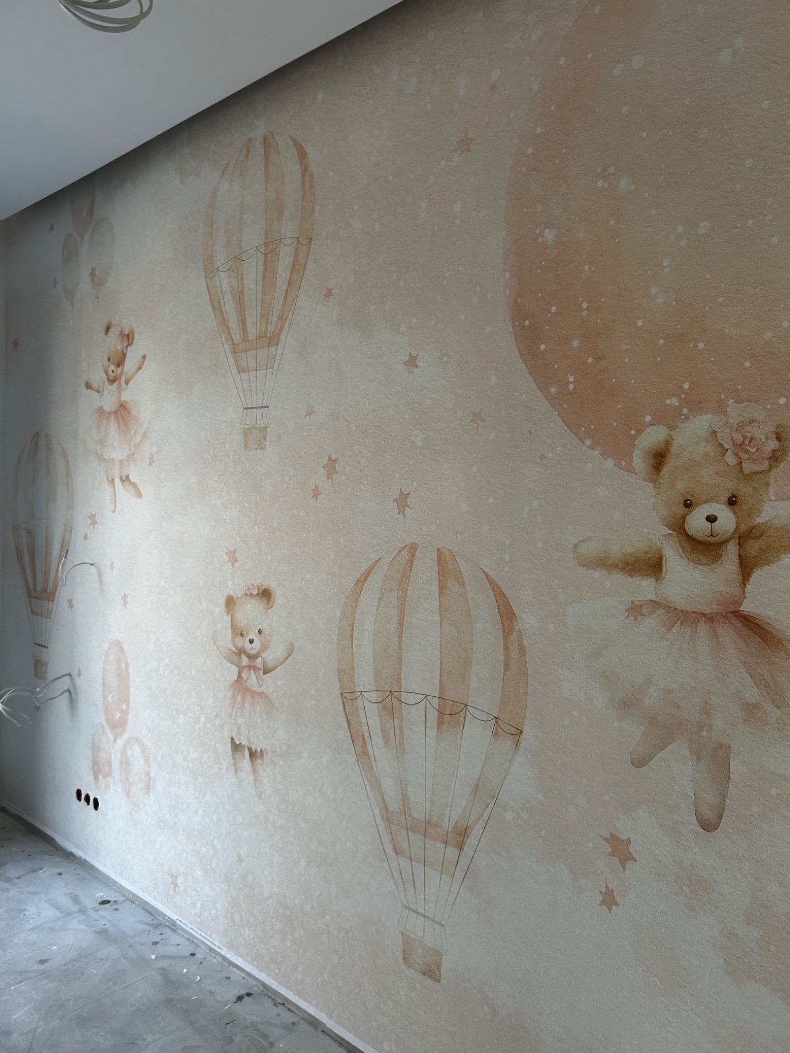 Ściana pokoju dziecięcego z tapetą przedstawiającą misie baletnice i balony na tle księżyca, widoczne gniazdka elektryczne i wystające kable.
