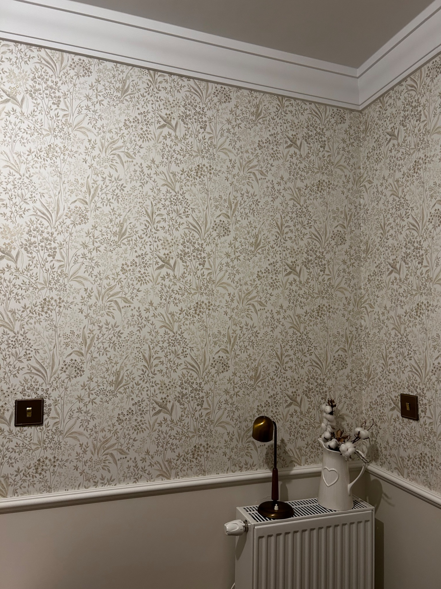 Wzorzysta tapeta w odcieniach beżu i brązu w pomieszczeniu w Łomży, uzupełniona listwami przypodłogowymi i sufitowymi, z widoczną lampką nocną i dzbankiem z bawełną na grzejniku.
