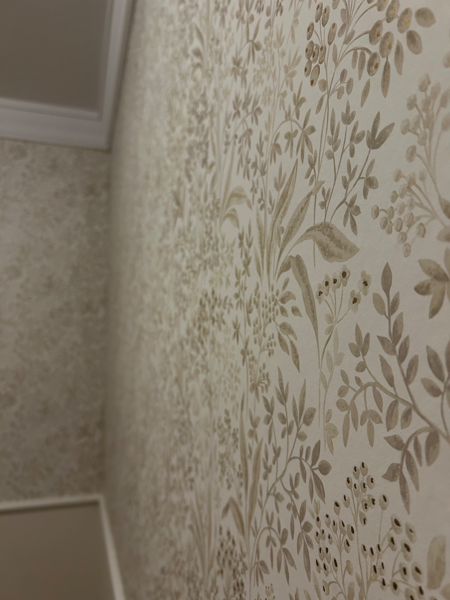 Ściana pokryta tapetą w delikatny, botaniczny wzór w odcieniach beżu i brązu, widoczny fragment sufitu z listwą dekoracyjną.