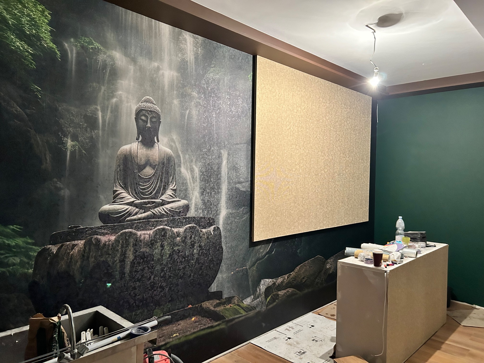 Tapeta z motywem Buddy i wodospadu obok fragmentu ściany z tapetą o drobnej fakturze, widoczne narzędzia i materiały malarskie.