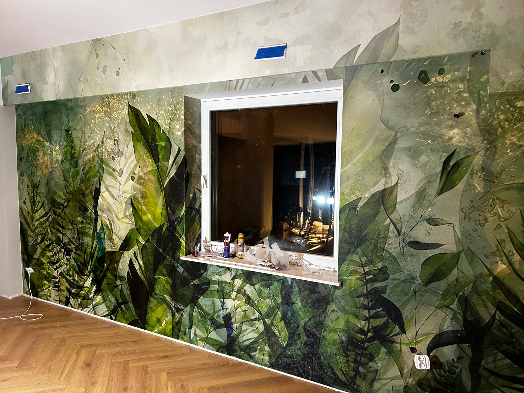 Fragment ściany z fototapetą o motywie tropikalnych liści w trakcie montażu, widoczne taśmy maskujące, przybory i preparaty na parapecie okna.