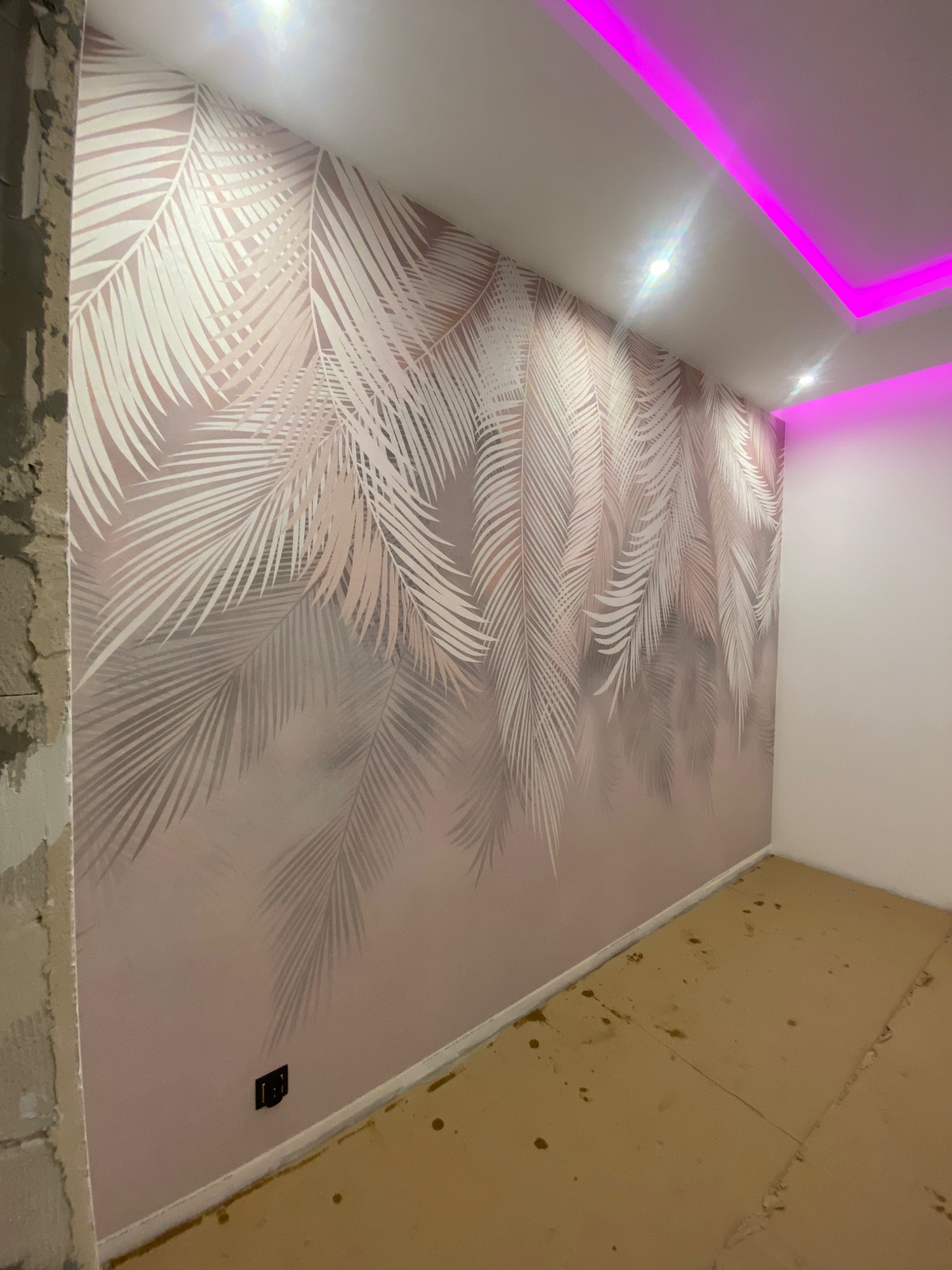 Ściana po nałożeniu tapety z motywem liści palmowych w odcieniach bieli i różu, oświetlona fioletowym światłem LED z sufitu. Fragment ściany w stanie surowym po lewej stronie.