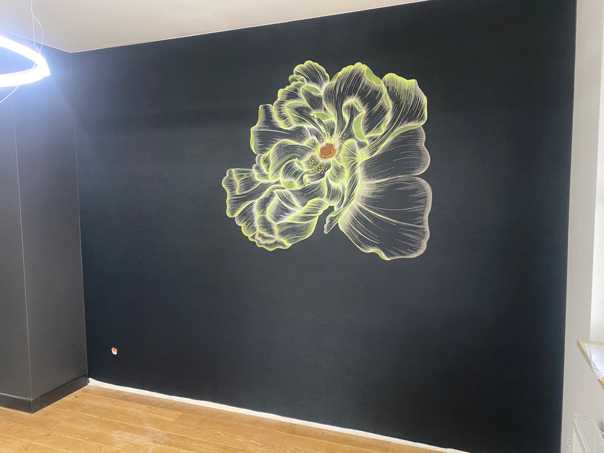Czarna ściana z ręcznie malowanym, świecącym motywem kwiatowym, widoczny fragment podłogi z paneli i nowoczesna lampa sufitowa.