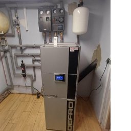 Mak-Tech - Szary piec na pellet marki Defro Evopell w kotłowni z widocznymi rurami instalacji grzewczej i zbiornikiem wyrównawczym. Na ścianie sterowniki i manometry.