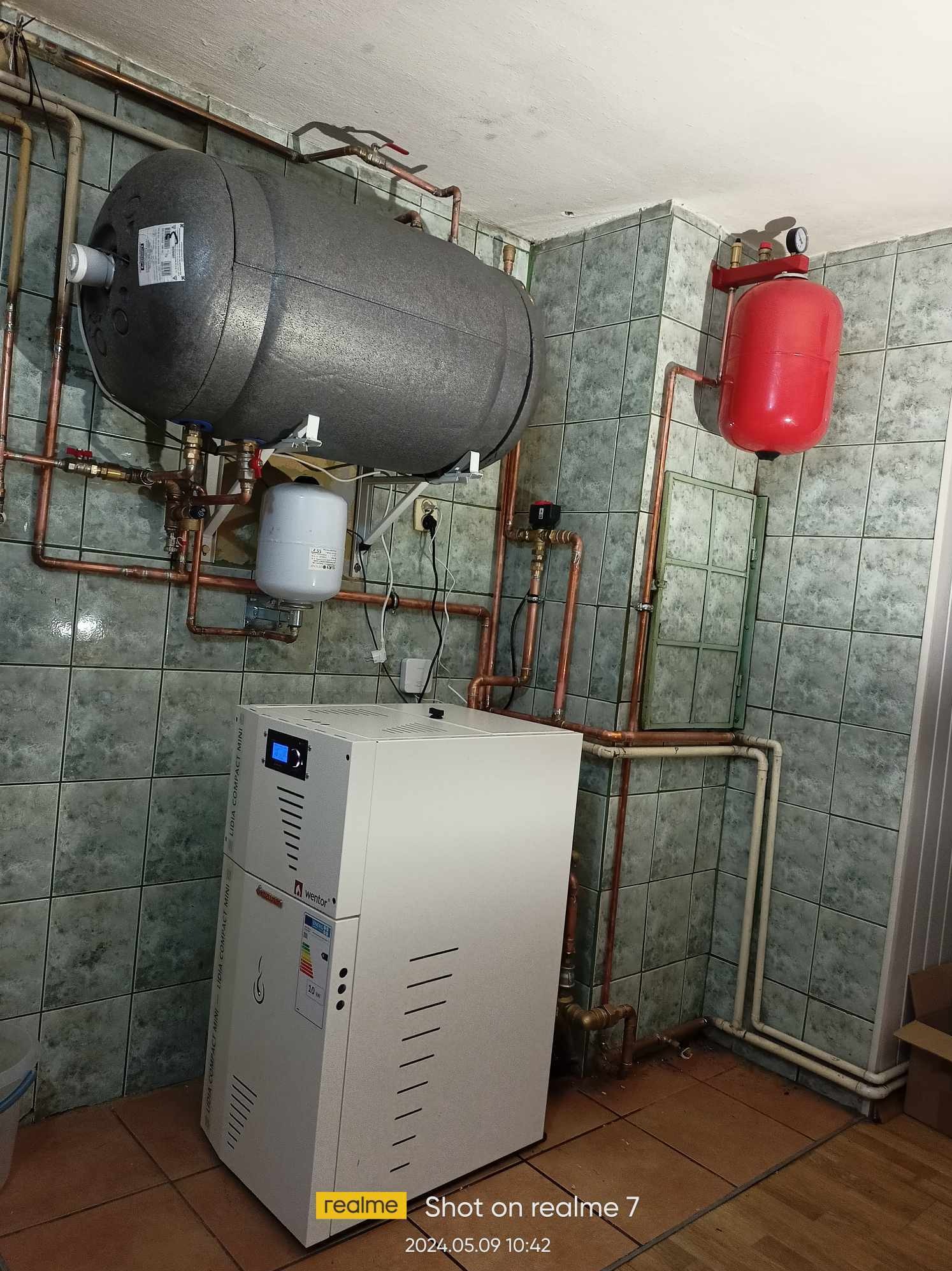 Nowoczesny, biały piec z wyświetlaczem i zasobnikiem ciepłej wody w pomieszczeniu z kafelkami, widoczne rury miedziane i elementy instalacji grzewczej.