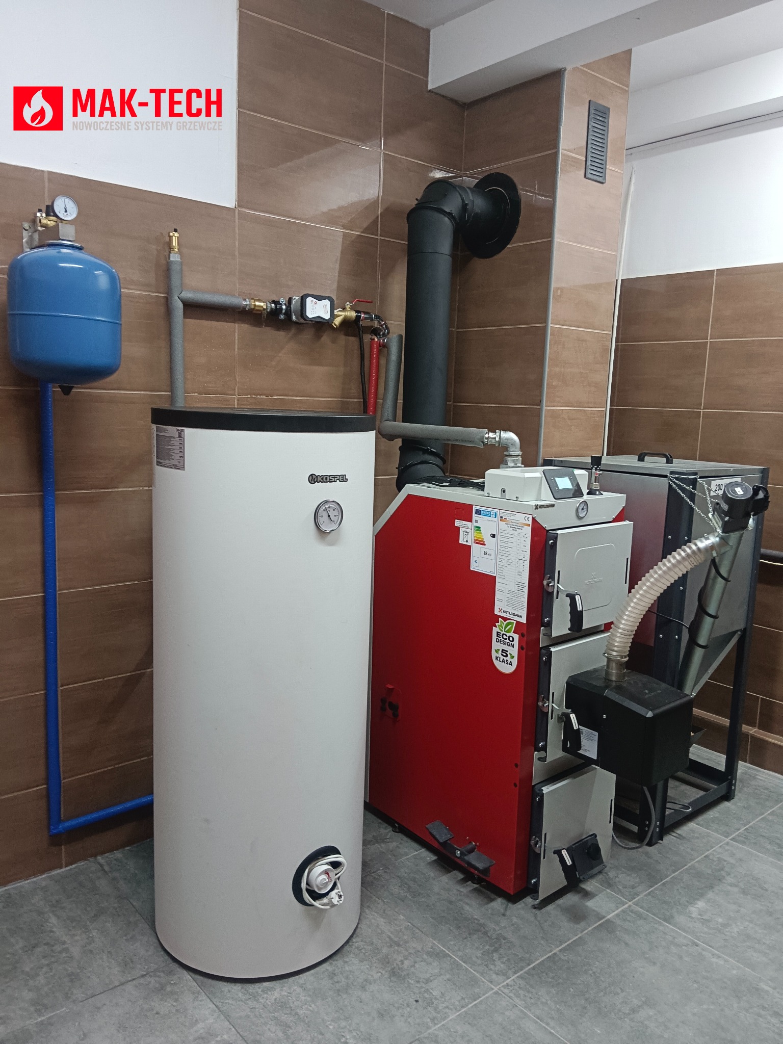 Instalacja kotła na paliwo stałe Kospel i zasobnika CWU w pomieszczeniu z brązowymi płytkami. Widoczny zbiornik wyrównawczy, rury, czarny komin i logo Mak-Tech.
